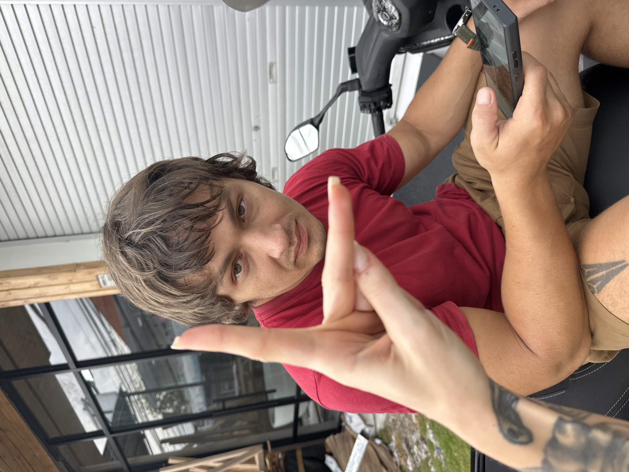 The image shows a man sitting on a scooter, holding a cell phone in his right hand. He is wearing a red shirt and khaki shorts. A hand with a tattoo on the arm is pointed at the camera. The setting appears to be outside, possibly on a porch or patio. The background includes corrugated metal siding, a window, and some vegetation. The picture was taken in Munggu, Indonesia.