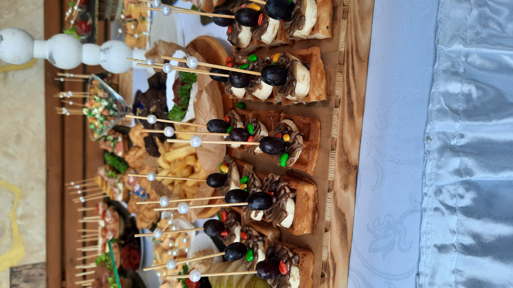 In Жодзішкаўскі сельскі Савет, Belarus, a catering setup displays a variety of appetizers and desserts. The foreground features a wooden board laden with bite-sized waffles topped with sliced bananas, chocolate drizzle, grapes, and colorful candy pieces, each secured with a decorative skewer. Behind these, a pile of french fries is visible in a paper-lined container. Further back, an assortment of other finger foods and salads are arranged on a table covered with a white tablecloth. The setting appears to be outdoors or in a space with a rustic wooden backdrop.