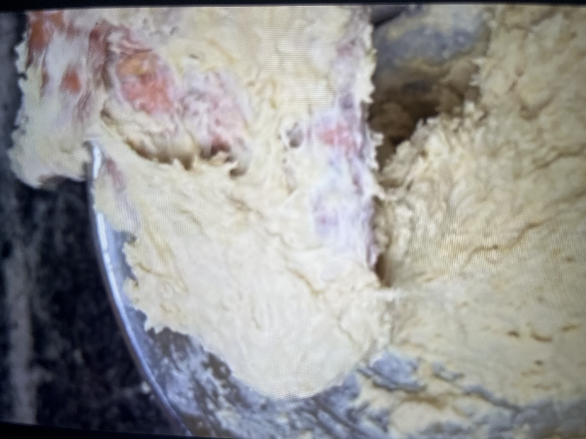 The image shows a close-up of a bowl of raw dough, likely for baking, with hints of reddish-pink ingredients mixed in. The texture is thick and creamy, with visible swirls from being mixed. The background is dark and out of focus, suggesting a kitchen environment. There are no people, actions, or interactions visible in the image. There is no indication of the city of Abuja, Nigeria in the image.