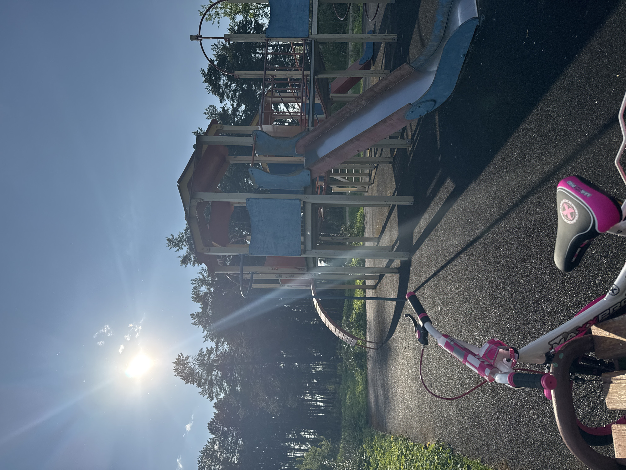 The image depicts a playground in Раздолье, Russia. The playground structure consists of wooden beams supporting a slide, climbing structures, and various platforms painted in blue and red. A portion of a child's bicycle with white and pink handlebars and a pink-accented seat is visible in the foreground. The playground is surrounded by trees, and the sun is shining brightly, casting long shadows. The ground is covered with a dark, textured material, likely rubber or asphalt.