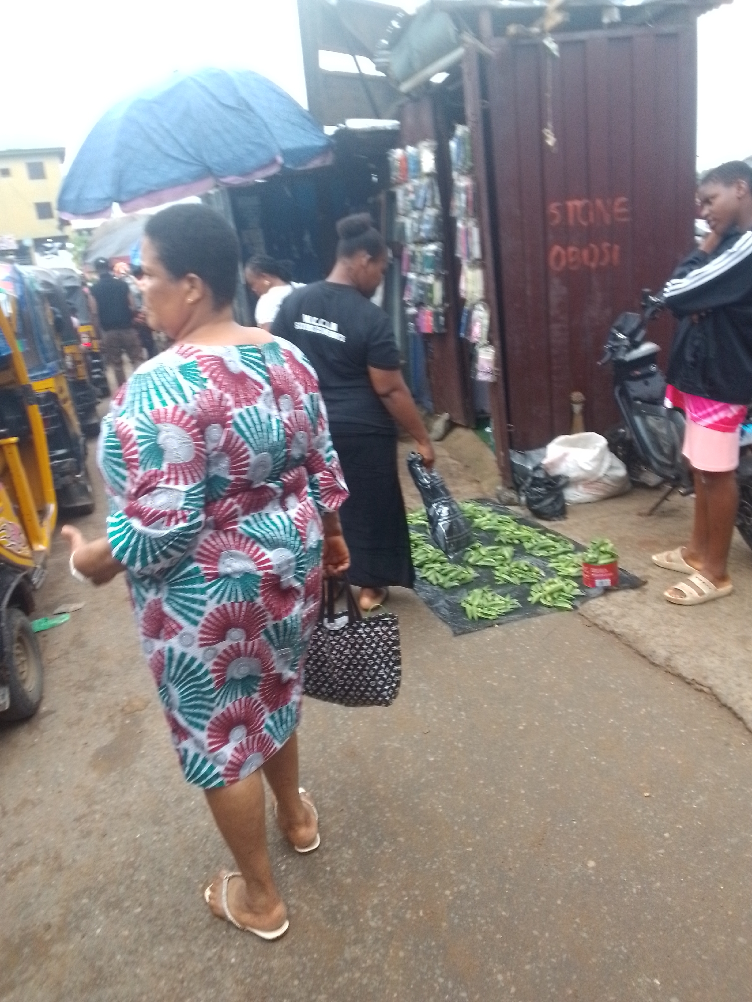 The image depicts a street market scene in Nkpor, Nigeria. In the foreground, a woman wearing a vibrantly patterned dress and white sandals walks away from the camera, carrying a patterned bag. To her left, a yellow motorized tricycle is partially visible. In the mid-ground, another woman in a black t-shirt and black skirt is seen standing near a stall. In front of her, green vegetables, possibly snap peas, are spread out on a black mat on the ground, with a few bags and a small red container also present. To the right, a person in a black jacket with white stripes and pink shorts stands near a motorcycle, looking towards the street. Behind the market stalls, a corrugated metal structure with various items, possibly phone cases, displayed on the left and red writing that reads "STONE OBOSI" on the right is visible. The ground is paved and appears to be a dirt or asphalt road. The sky is overcast, suggesting cloudy weather.