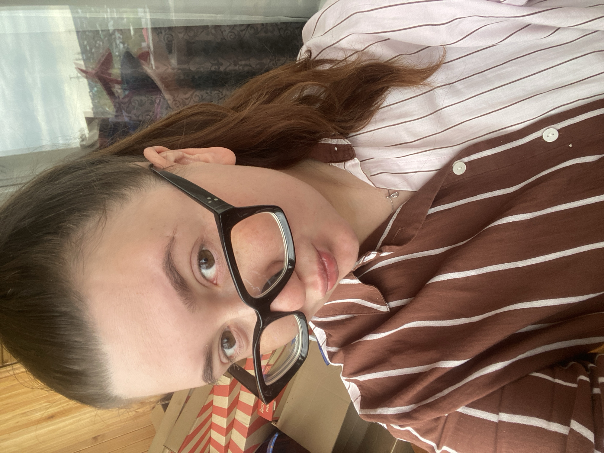 A woman is facing the camera, wearing black glasses and a striped shirt that appears to be a mix of brown and pink with white stripes. Her hair is brown and pulled back. The background is a cluttered scene with boxes and a window reflecting an outdoor scene. No information allows to determine if the image was taken in the city of Chaikovsky, Russia.