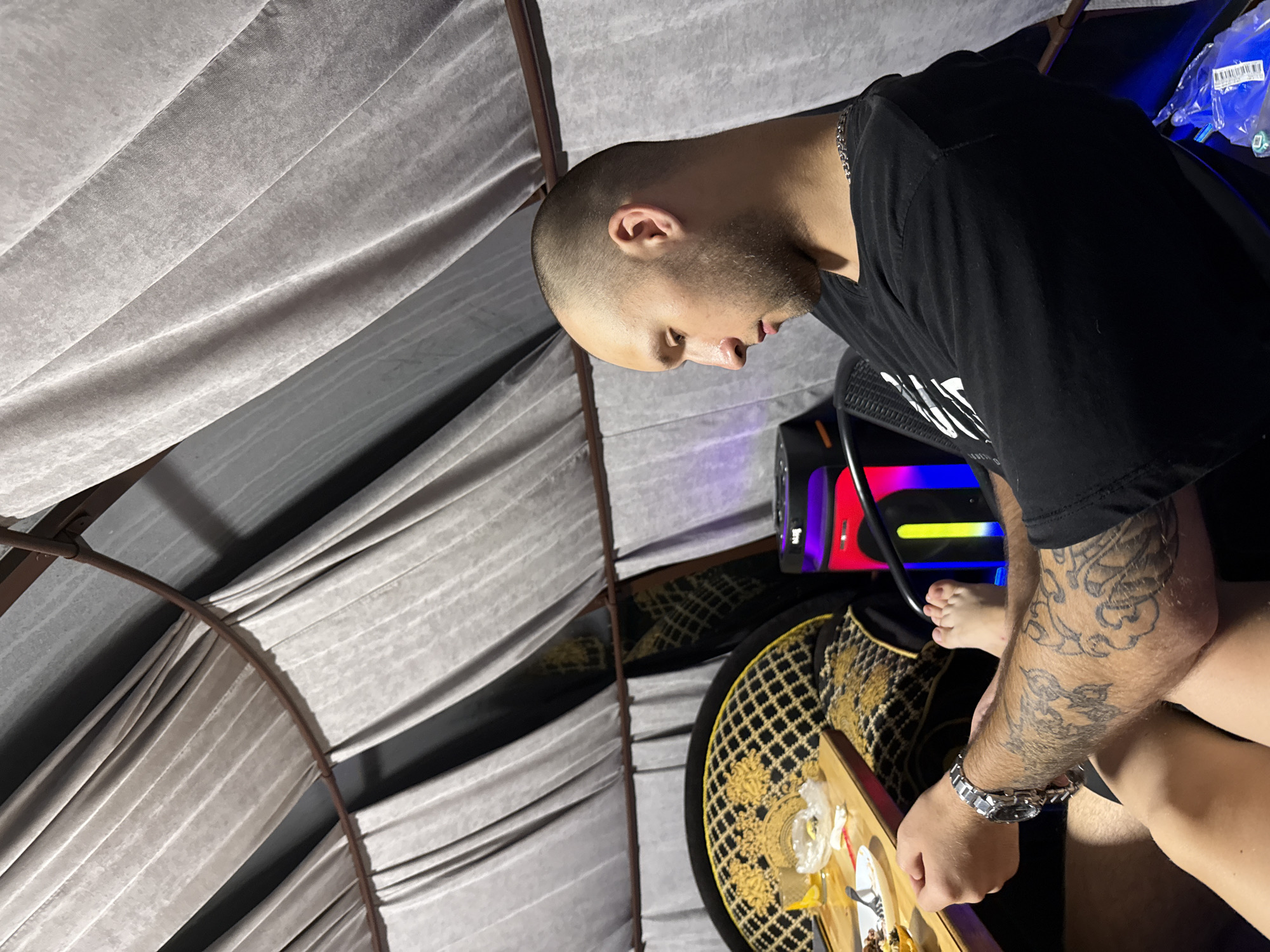 In an enclosed space with draped fabric walls, a man with a shaved head and a beard sits in profile, looking down. He is wearing a black t-shirt and a silver watch on his left wrist. His left arm and forearm are covered in tattoos. In front of him, a wooden table holds plates of food, a glass with a yellow drink, and a small plastic bag. To his right, a black speaker with colorful LED lights is visible. A black mesh chair is behind him. His bare feet are visible. The overall impression is of a relaxed or contemplative moment within a somewhat decorated indoor setting.