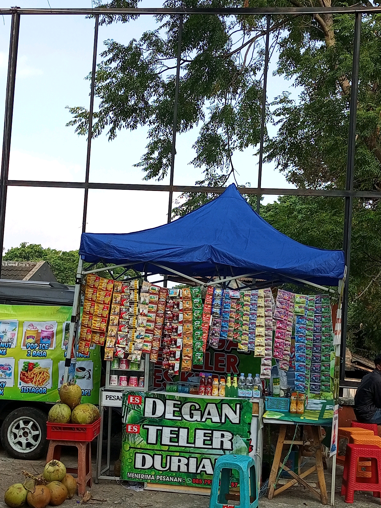 In Batu, Indonesia, an outdoor street vendor stall operates under a blue tarp, displaying a variety of packaged snacks and beverages. The stall is set up on a paved surface, likely a sidewalk or roadside, with a green and white mobile cart visible to the left. Coconuts are prominently displayed, some in a red crate on a small red stool, and others scattered on the ground in the foreground. The stall's merchandise includes rows of colorful snack packets hanging from racks, and bottled drinks arranged on shelves. A sign on the cart advertises "Pop Ice" and "Sistacor." Further back, a larger sign in green lettering reads "DEGAN TELER DURIAN" with a phone number at the bottom, indicating a beverage or food service, likely selling coconut and durian-based drinks. A person in a black shirt is seated on a red stool to the right of the stall. The scene is framed by a metal structure with glass panels reflecting trees and the sky, suggesting the stall is positioned in front of a building or a large glass-enclosed area. The overall environment appears to be an urban street with trees and clear sky.