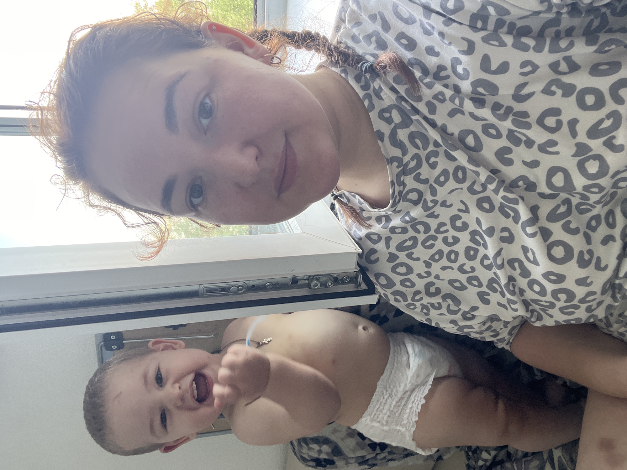 A woman and a baby are captured in a selfie, seemingly taken indoors. The woman, with auburn hair styled in braids and wearing a leopard-print shirt, is positioned in the foreground, her face close to the camera. She has noticeable freckles and is wearing a hoop earring in her visible ear.
To her left and slightly behind, the baby, wearing only a diaper, is looking towards the camera with an open mouth and an outstretched hand. The baby has a small mark on its forehead.
The background suggests a domestic setting. A window with a view of greenery is visible to the right. Part of a door or window frame with visible hardware is also present. The overall lighting indicates it's daytime, possibly with sunlight filtering in. There is no identifiable signage or landmarks to indicate the specific location within Chelyabinsk, Russia. The scene is informal and candid.