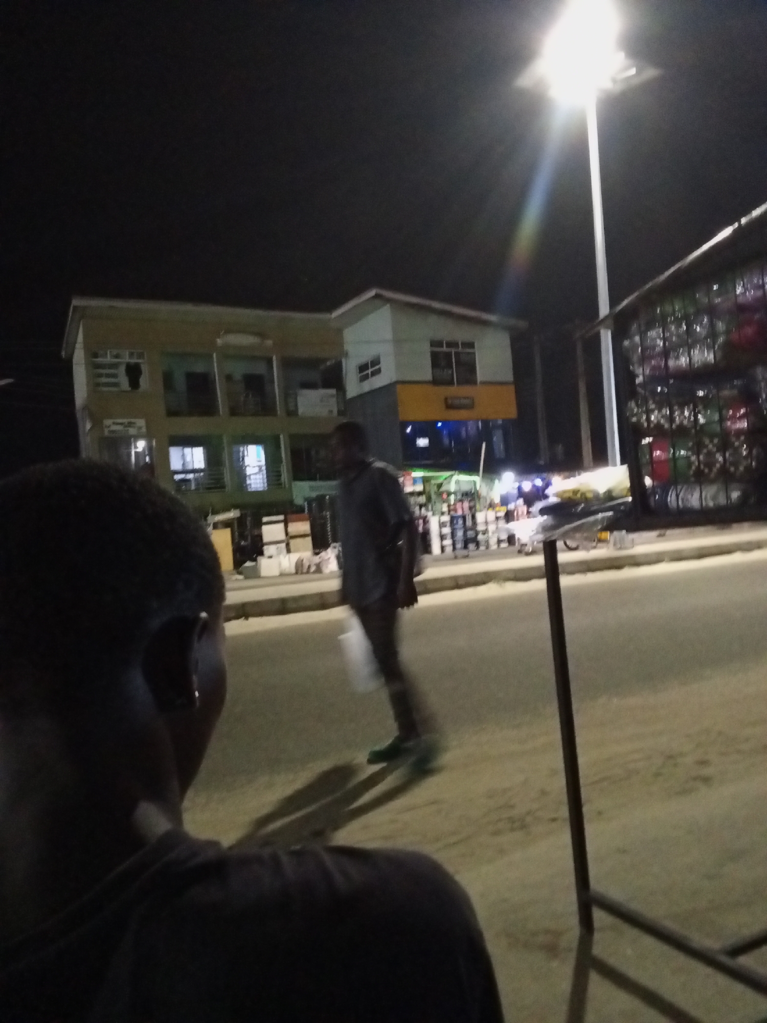 The image depicts a nighttime street scene in an urban or semi-urban area, likely in Ado, Nigeria. In the immediate foreground, the back of a person's head and left shoulder are visible, indicating the perspective of an observer; a small, reflective earring is present on the ear.
A tall, bright street light on a white pole dominates the upper right, casting strong illumination over the scene and producing a noticeable lens flare with a subtle rainbow effect. Below the light, on the right side of the street, a shop or stall is visible, featuring shelves packed with numerous items, possibly bottles, containers, or various goods.
Across the paved street, a man is captured in motion, walking from left to right. He wears a dark shirt and trousers, and appears to be carrying a light-colored object. His shadow stretches long behind him on the sandy or dirt shoulder of the road.
In the background, on the far side of the street, are multi-story buildings. The building on the left has a light-colored exterior with several windows, some emitting light from within. A small yellow sign is visible on this building, but any text on it is illegible due to distance and blur. Another building with a white section and dark windows is partially visible further right. The sky is dark, confirming the night setting. A metallic structure, possibly a stand or railing, is present in the lower right foreground.