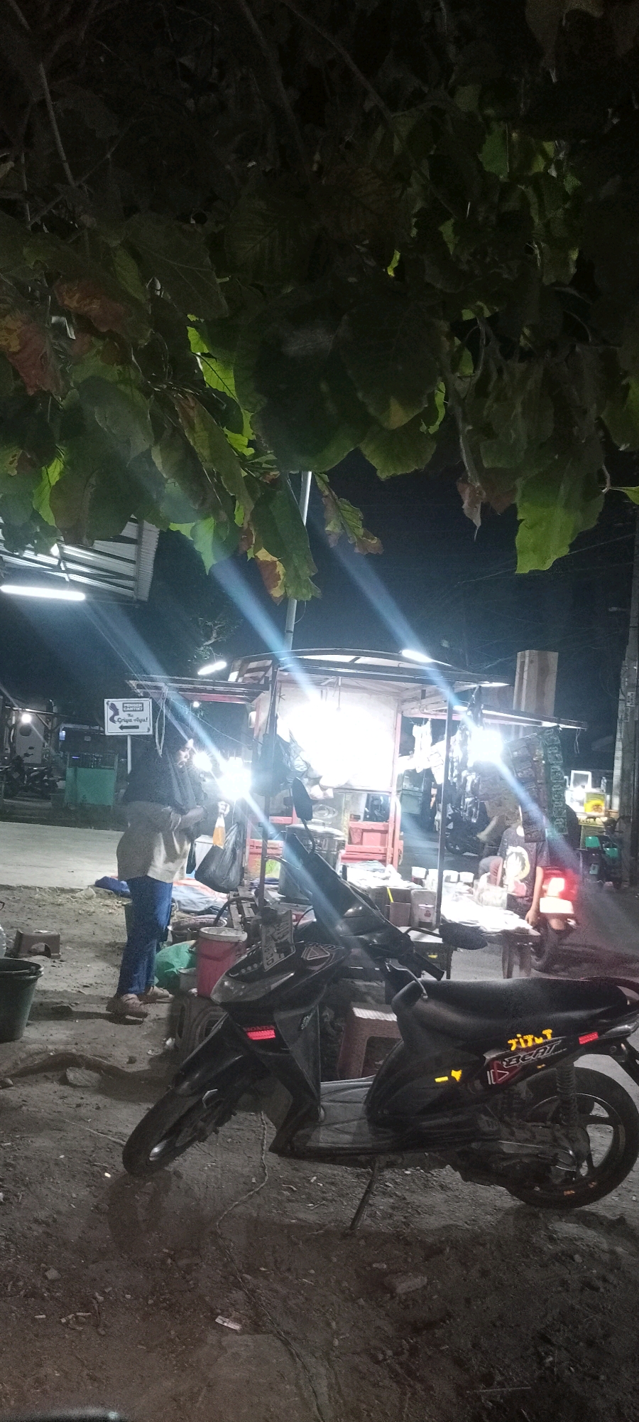 The image depicts a street-side scene at night, likely in Bendotretek, Indonesia, under a canopy of leaves. In the foreground, a dark-colored motorcycle is parked, its details partially obscured by shadows. A woman wearing a dark hijab and a light-colored long-sleeved shirt and blue pants stands to the left of a food stall. She is holding a clear plastic bag. The food stall, illuminated by bright lights, appears to be set up on a cart with various items displayed. To the left of the woman, a green bucket is visible. The ground is uneven and appears to be a dirt or gravel surface. In the background, more stalls or buildings are faintly visible, along with some parked vehicles. The overall atmosphere suggests a local market or a small roadside eatery operating in the evening.