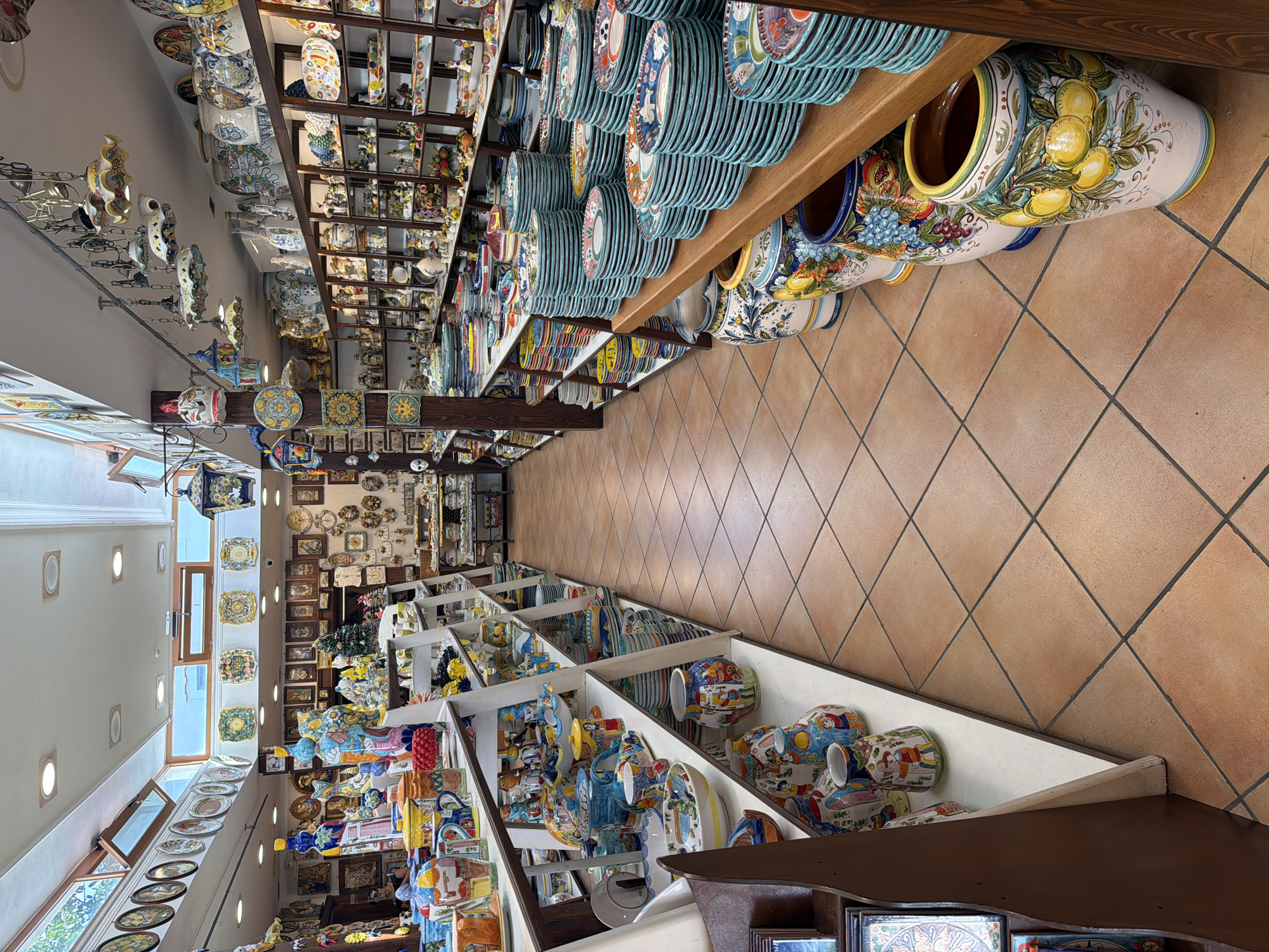 The image depicts the interior of a shop filled with artisanal ceramic products, likely located in Ischia, Italy. The space is narrow and deep, with shelves lining both sides of a central aisle. The shelves are laden with stacks of ceramic plates, vases, and decorative objects, all featuring vibrant, hand-painted designs. The floor is tiled in a terracotta color. The lighting is bright, suggesting daylight. No people are visible in the immediate frame. The overall impression is of a retail space dedicated to local crafts and ceramics.