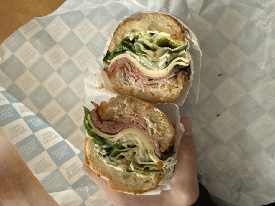 A large sandwich, cut in half, is centrally positioned and held by a human left hand. The sandwich is constructed on a long, light-brown bread roll, featuring layered contents including thinly sliced pinkish-red deli meat, light yellow cheese, green leaf lettuce, and a visible creamy white sauce or dressing. Darker elements, possibly olives or a specific spread, are visible at the bottom of the filling. The sandwich rests on white butcher paper with a light blue checkerboard pattern. The pattern incorporates repeated text "COURT STREET GROCERS," indicating the sandwich's origin from Court Street Grocers, an establishment located in Brooklyn, New York City, United States of America. A light brown surface, likely a wooden table or countertop, is partially visible beneath the paper in the upper right background.