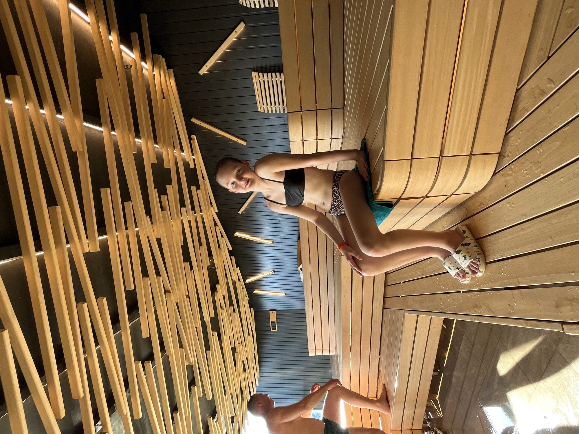 The image shows the interior of a sauna or steam room. A girl in a black bikini top and animal print bottoms is sitting on a wooden bench with a towel behind her. She is smiling and looking at the camera. A man is sitting on a lower bench in the background, facing away from the camera. The walls and benches of the sauna are made of light-colored wood. The ceiling is decorated with irregularly placed wooden slats and lights. The image was taken in a sauna, but the specific location of Votkinsk, Russia, is not definitively identifiable from the visual information in the photograph. The environment is a sauna or steam room, with wooden features and bathing attire worn by the people present.