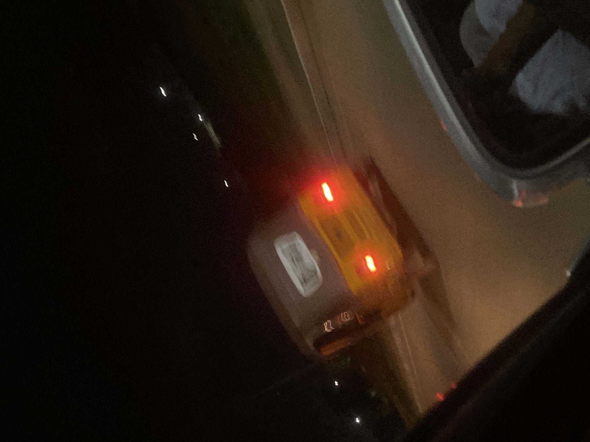 The image, taken at night, shows the interior of a car. The car's dome light is on, emitting a yellowish glow with two visible red lights. A portion of the car's side mirror and a blurred reflection of a person's legs, covered in light-colored fabric, are visible. Outside the car window, the dark background suggests the setting is urban. The series of lights, ascending diagonally, suggest that this picture was captured in the city of Abuja, Nigeria.