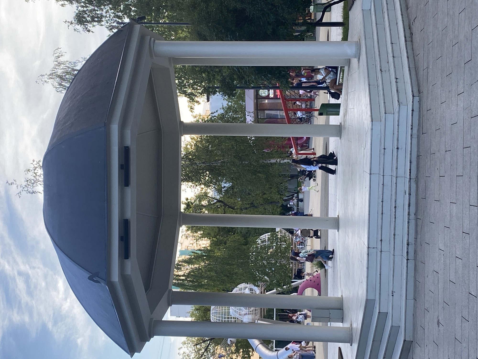 The image captures a large, octagonal gazebo with a dark, domed roof and white pillars, set on a raised white platform with multiple steps. It is situated in a public park area, likely a children's playground, given the presence of colorful play structures, including a silver slide and white spherical climbing frames, in the background. Several people are visible in the park, including a woman walking with a stroller, a man in a black t-shirt, and a woman sitting on a bench, looking at her phone. The environment is green and leafy, with trees and buildings visible beyond the park. The overall impression is one of a lively urban park during daylight hours, with a slightly overcast sky. Based on the architectural style of the gazebo and the surrounding urban park elements, this scene is consistent with a location within the city of Moscow, Russia.