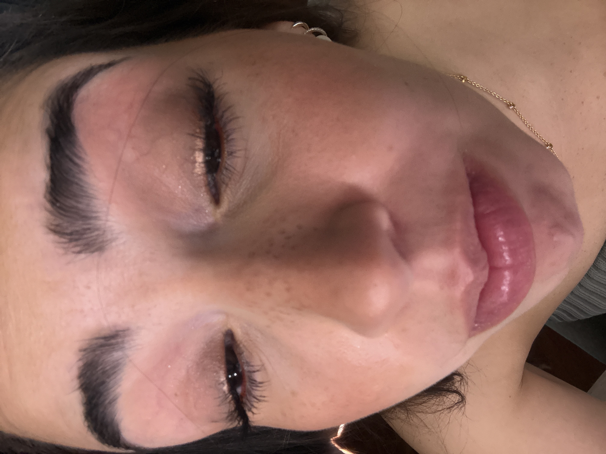 A close-up, slightly angled shot of a woman's face. She has dark eyebrows, closed eyes, and full lips. She also has freckles. A gold necklace is visible on her neck, and an earring is seen in her ear. The background is slightly blurred, with hints of fabric and hair. The location cannot be determined from the image.