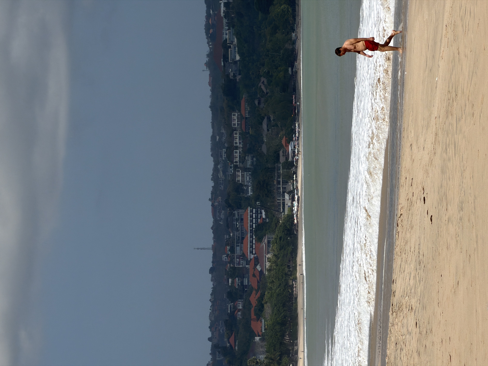 The image captures a segment of Kuta Beach in Indonesia under a cloudy sky. In the foreground, a man in red swimming trunks walks along the wet sand near the edge of the ocean. He is bare-chested and appears to be walking away from the camera, towards the right side of the frame. The ocean displays a gentle surf with white-capped waves lapping at the shore. In the mid-ground, the sandy beach stretches back, dotted with what appear to be beach chairs and umbrellas, suggesting a developed tourist area. Further in the background, a lush green hillside is covered with buildings, their red-tiled roofs characteristic of Balinese architecture, and some taller structures and a telecommunications tower are visible on the higher slopes. The distant sky is overcast with dark, heavy clouds, creating a somber mood.