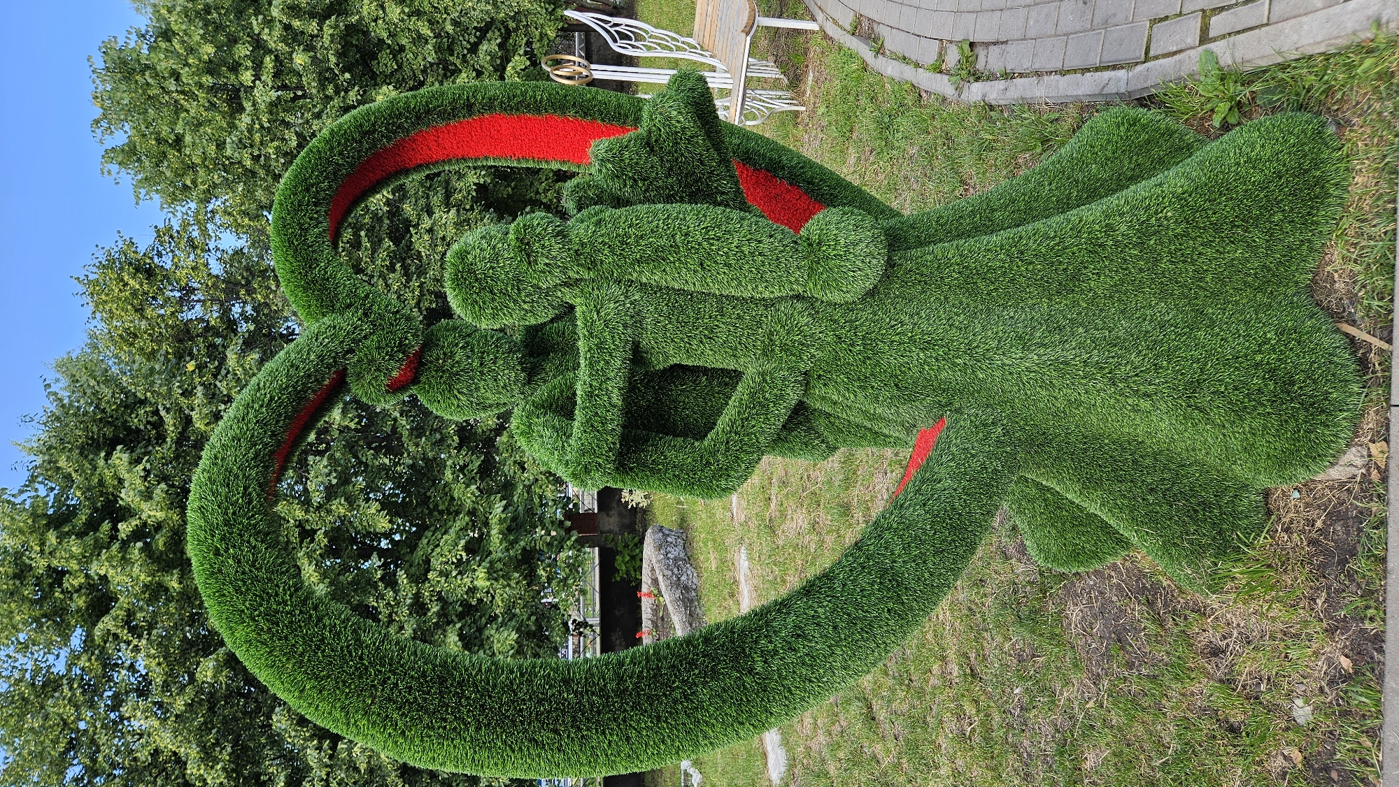 The image depicts a heart-shaped topiary sculpture in Klyavlino, Russia. The sculpture is made of green artificial grass and features two figures embracing within the heart. Red material lines the inside of the heart shape, contrasting with the green of the topiary. The figures are also covered in the green artificial grass, with their arms wrapped around each other. The sculpture is situated outdoors on a grassy area, with trees and a clear blue sky visible in the background. A paved walkway runs along the right side of the sculpture. In the distance, a park bench with decorative white wings can be seen.