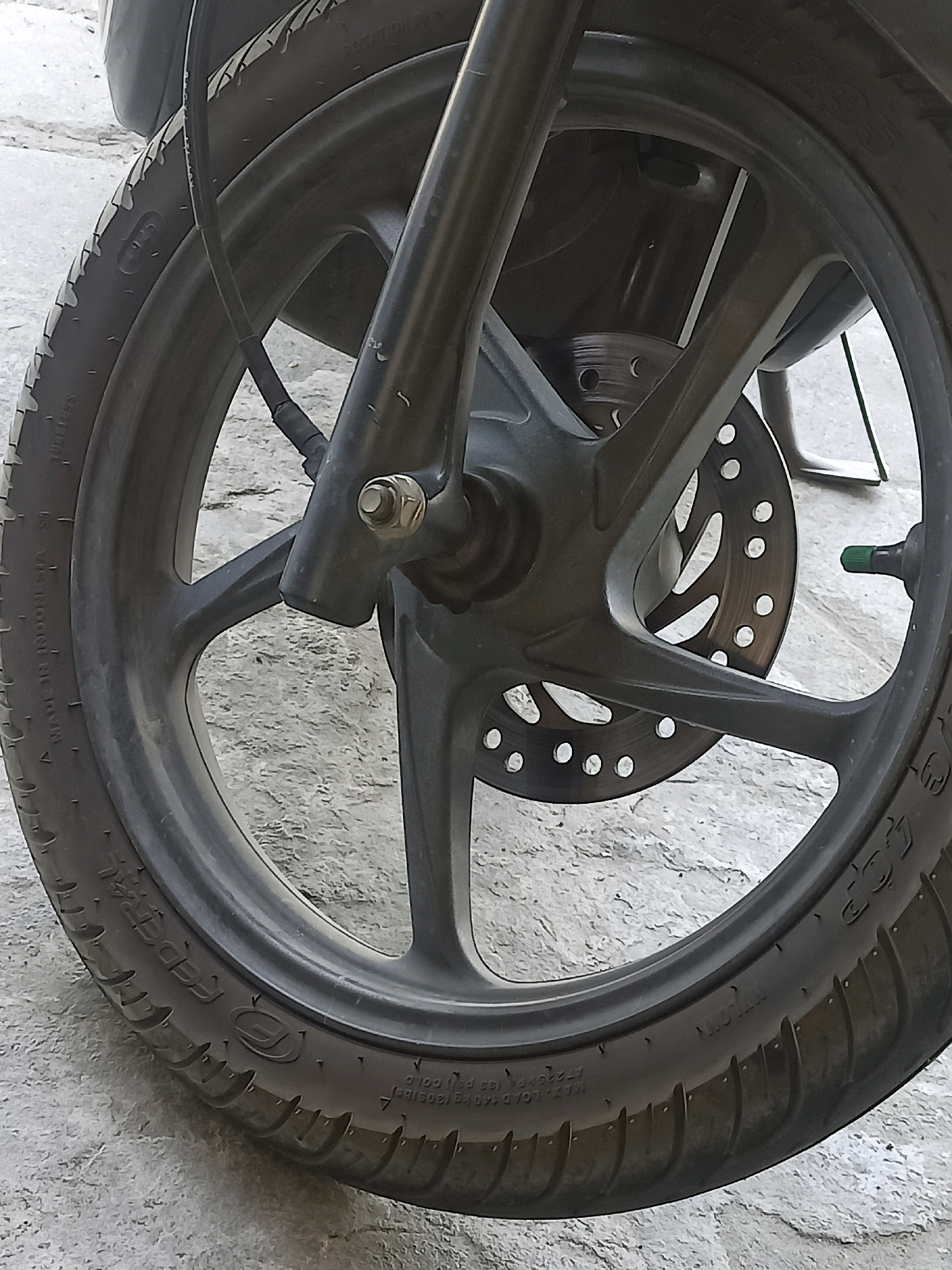 The image shows a close-up of a motorcycle's front wheel, specifically focusing on the tire, rim, brake disc, and part of the front suspension fork. The wheel is black with a five-spoke alloy rim. The tire is black with visible tread patterns and markings indicating it is a "FEDERAL" brand tire and is "TUBELESS." There are also pressure and load specifications visible on the sidewall, reading "MAX. LOAD 400 KGS (130PSI)." The brake disc is a circular metal plate with multiple perforations. The front suspension fork is black and robust, with a visible bolt and nut assembly. A green valve stem cap is present on the wheel. The background consists of a rough, light-colored concrete surface, likely a road or pavement. The scene appears to be outdoors in what could be a street or alleyway within the city of Gabungan, Indonesia, given the context provided. There are no people or animals visible in the image, and no apparent actions or interactions are occurring. The overall lighting suggests it is daytime.