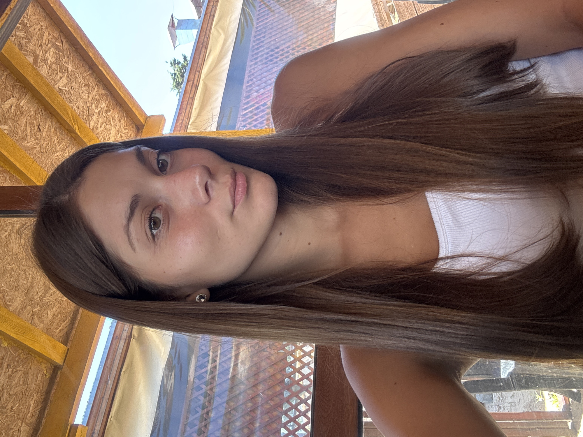 A young woman with long, straight brown hair and brown eyes is photographed from the chest up in a selfie. She is wearing a white tank top and a small stud earring in her left ear. She has a light tan complexion with some freckles on her nose and cheeks. The background suggests an outdoor setting with a wooden roof structure above and a blurred background of what appears to be outdoor seating with a lattice-like screen and some greenery. The lighting indicates it is daytime, likely outdoors. The image does not provide specific details about the city of Sozopol, Bulgaria.