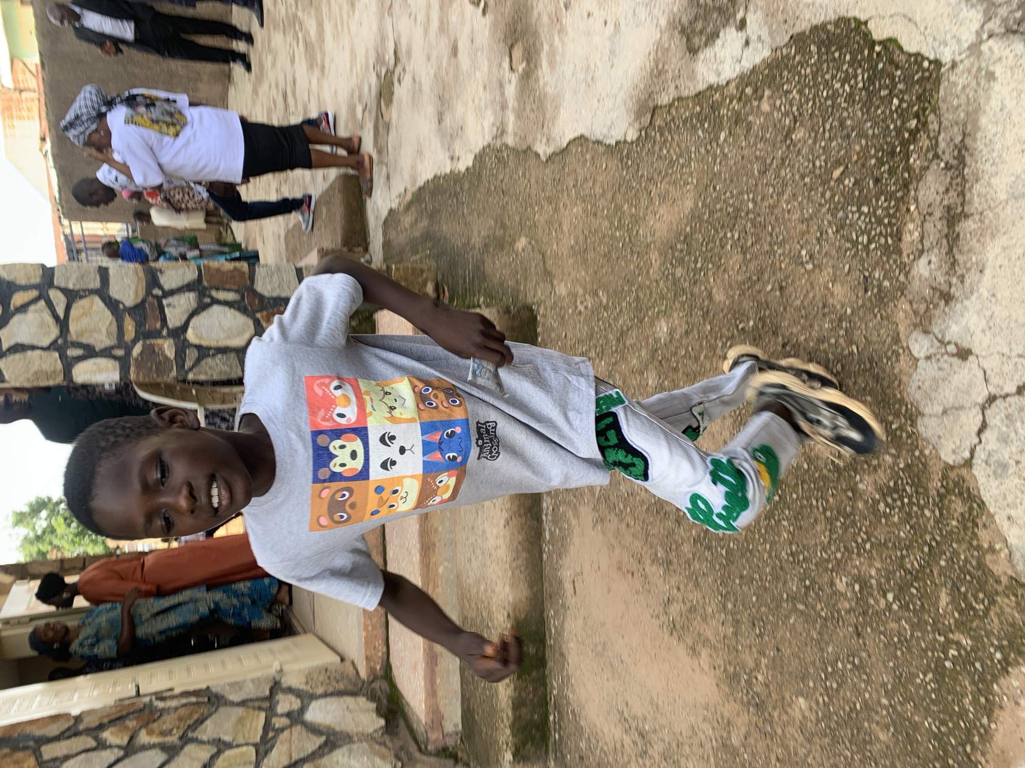 A young Black boy runs across a textured, dirt and concrete path in an outdoor, urban setting. He is captured in motion, his left leg extended and his right leg bent, suggesting forward momentum. The boy wears a light grey t-shirt featuring a grid of cartoon animal faces, a popular video game motif. His pants are light-colored with a repeating green design of an alligator or crocodile, and he wears dark and light-colored athletic shoes. In his right hand, he holds a few folded paper bills.
Behind the boy, a stone wall forms the exterior of a building. To the left, an open doorway reveals a glimpse of individuals inside, including a woman in a blue and white patterned dress and another woman in an orange garment. Further in the background, other people are visible, including a man in a suit. The environment suggests a residential or communal area within the city of Jos, Nigeria, characterized by its masonry construction, uneven ground, and the presence of multiple individuals engaged in various activities. The lighting indicates it is daytime.
