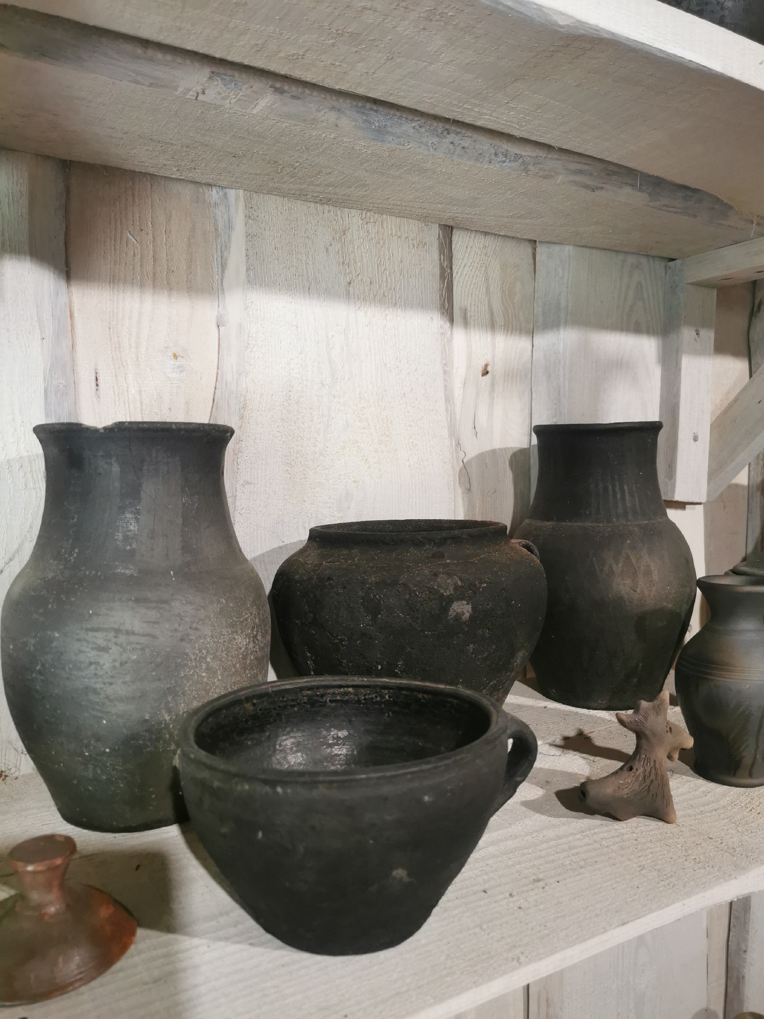 An arrangement of rustic, dark-colored ceramic pots and a small clay figurine are displayed on a light-colored wooden shelf. The pots vary in size and shape, with some having wide mouths and others being more elongated. The figurine appears to be an animalistic shape. The background consists of vertical wooden planks. The scene is indoors, likely in a rural setting in or around Шчарчоўскі сельскі Савет, Belarus, suggesting a historical or cultural display.