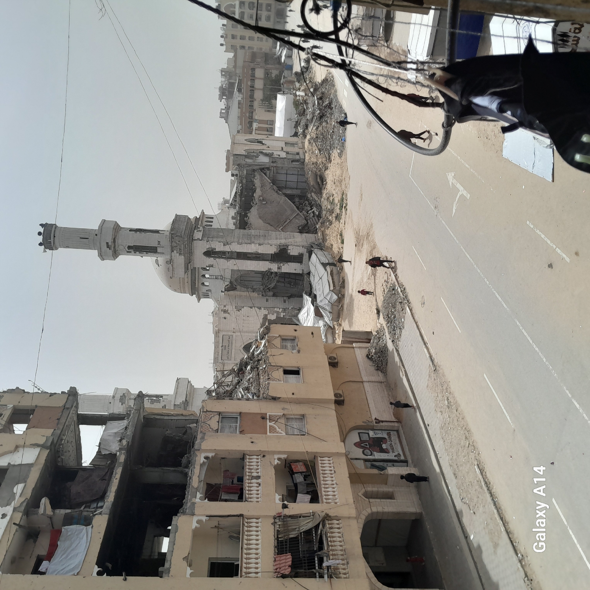 The image shows a street in Khan Yunis, Palestinian Territory, visibly damaged from conflict. Buildings are heavily damaged, with some partially collapsed. Debris litters the street. People are present, walking along the street. A mosque with a prominent minaret is damaged but still standing. A road with white markings runs through the scene. Overheard wires are present in the foreground. A sign for "Yaseen Games" is visible on a building.