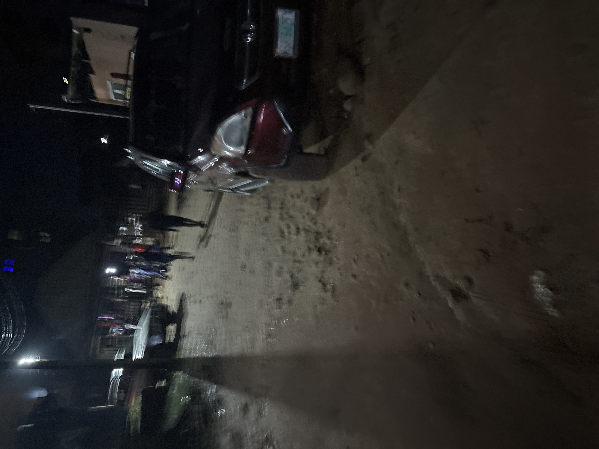 The image depicts a dimly lit, muddy street scene at night in Onitsha, Nigeria. A dark-colored SUV is parked on the right side of the road, facing away from the camera. Its headlights are off, and reflections are visible on its surface. In the background, several figures, possibly pedestrians, are walking along the street. One individual in a blue shirt is clearly visible. The street appears to be unpaved and wet, with puddles of water and scattered debris. A light post illuminates part of the scene, casting a long shadow across the muddy ground. In the distance, structures that resemble shops or buildings can be seen, with some lights illuminating them. The overall atmosphere is dark and somewhat chaotic, suggestive of a busy, informal urban environment at night.