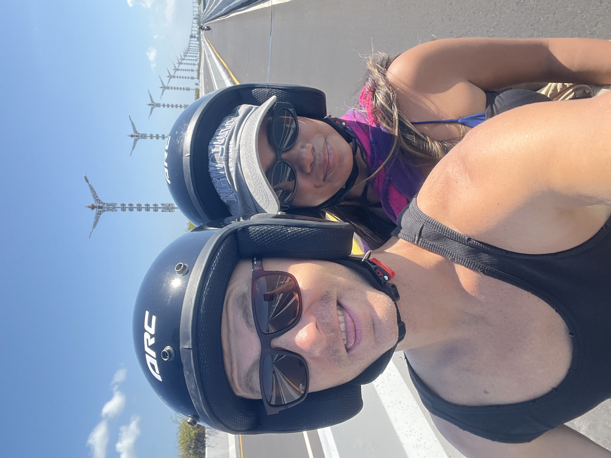 A selfie taken by a man and a woman riding a scooter on a bridge with palm-tree-shaped streetlights in the background. The man in the foreground wears a black helmet with "ARC" written on it, sunglasses, and a black tank top. He is sticking his tongue out. The woman behind him wears a gray baseball cap with "NEW YORK" embroidered on it, sunglasses, and a purple and pink scarf. They are on a road with white lines and a concrete barrier. The sky is clear and blue. The location is the Bali Mandara Toll Road in Kuta Selatan, Indonesia.