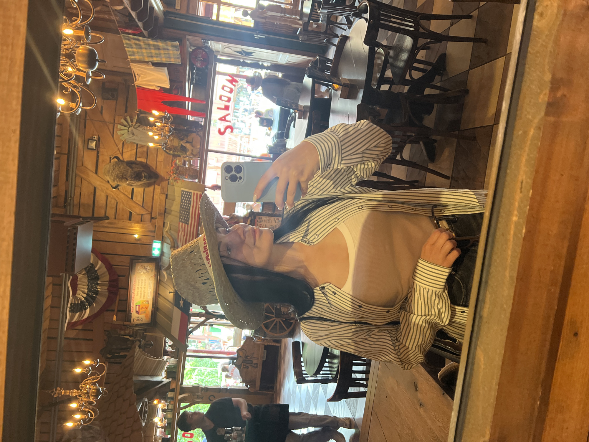 The image depicts a reflection in a mirror of a woman in a straw cowboy hat, a striped button-up shirt, and a white tank top, taking a selfie with her phone. She is seated at a wooden table in a restaurant with a Western theme, potentially the Saloon restaurant in Collendoorn, Netherlands, based on the visible "SALOON" signage. The background of the reflection reveals the restaurant's interior, which features wooden paneling, chandeliers, mounted animal heads, and American flags. Several tables and chairs are visible, as are other patrons. The woman's hands are visible, one holding the phone and the other resting on her lap, adjusting her belt. The overall scene suggests a casual dining or entertainment environment with a distinct Wild West aesthetic.