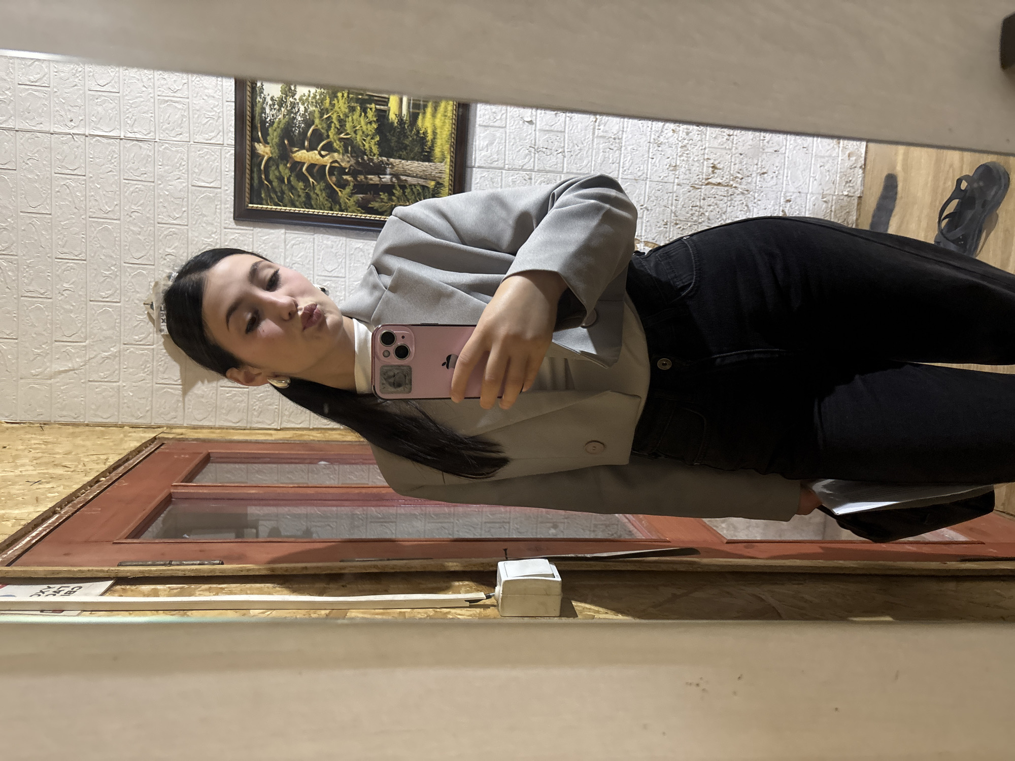 A young woman with dark hair, wearing a light gray cropped blazer over a white turtleneck and black pants, takes a mirror selfie. She has a light-colored headband on her head and large hoop earrings. She is holding a pink smartphone in her right hand, and a black bag is held in her left hand. The background appears to be an indoor space with textured white brick wallpaper on the right and a wooden wall with a red-framed door on the left. A framed painting of a tree is visible on the brick wall. There is a light switch on the wooden wall and a pair of black sandals on the floor reflected in the mirror. The scene suggests a casual, personal moment captured in a home or an informal setting. The specific location in the city of Fokino, Russia, is not directly identifiable from the visual elements of the image.
