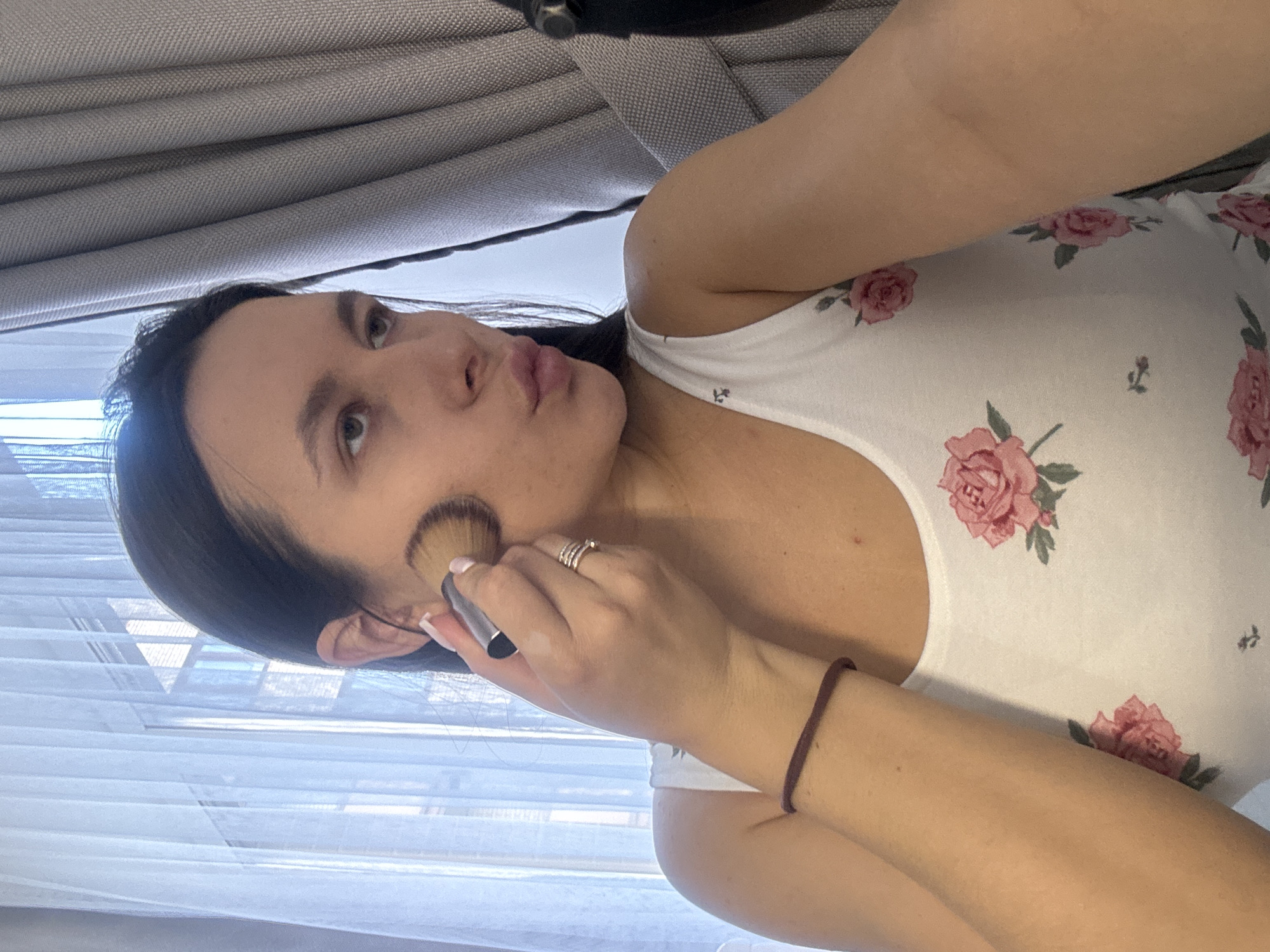 A woman is applying makeup. She is holding a brush in her right hand and applying it to her cheek. She is wearing a white tank top with a floral pattern. She has dark hair and light skin. The background is a window with blinds and a gray cushion. The location is possibly in Odintsovo, Russia, based on the user's request.