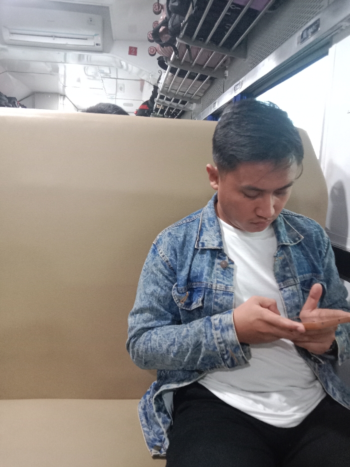 The image shows a young man wearing a blue denim jacket and white t-shirt sitting on a beige seat. He is looking down at a mobile phone in his hands. The man has short black hair. Overhead luggage racks are visible. The scene is likely taking place inside a train, likely in the city of Kedungjati, Indonesia at the provided coordinates. There is no visible evidence of any other people present in this scene. The ambient lighting is bright and appears to be coming from the overhead fluorescent lights in the train car. The windows to the left and right of the man are not visible in the image.