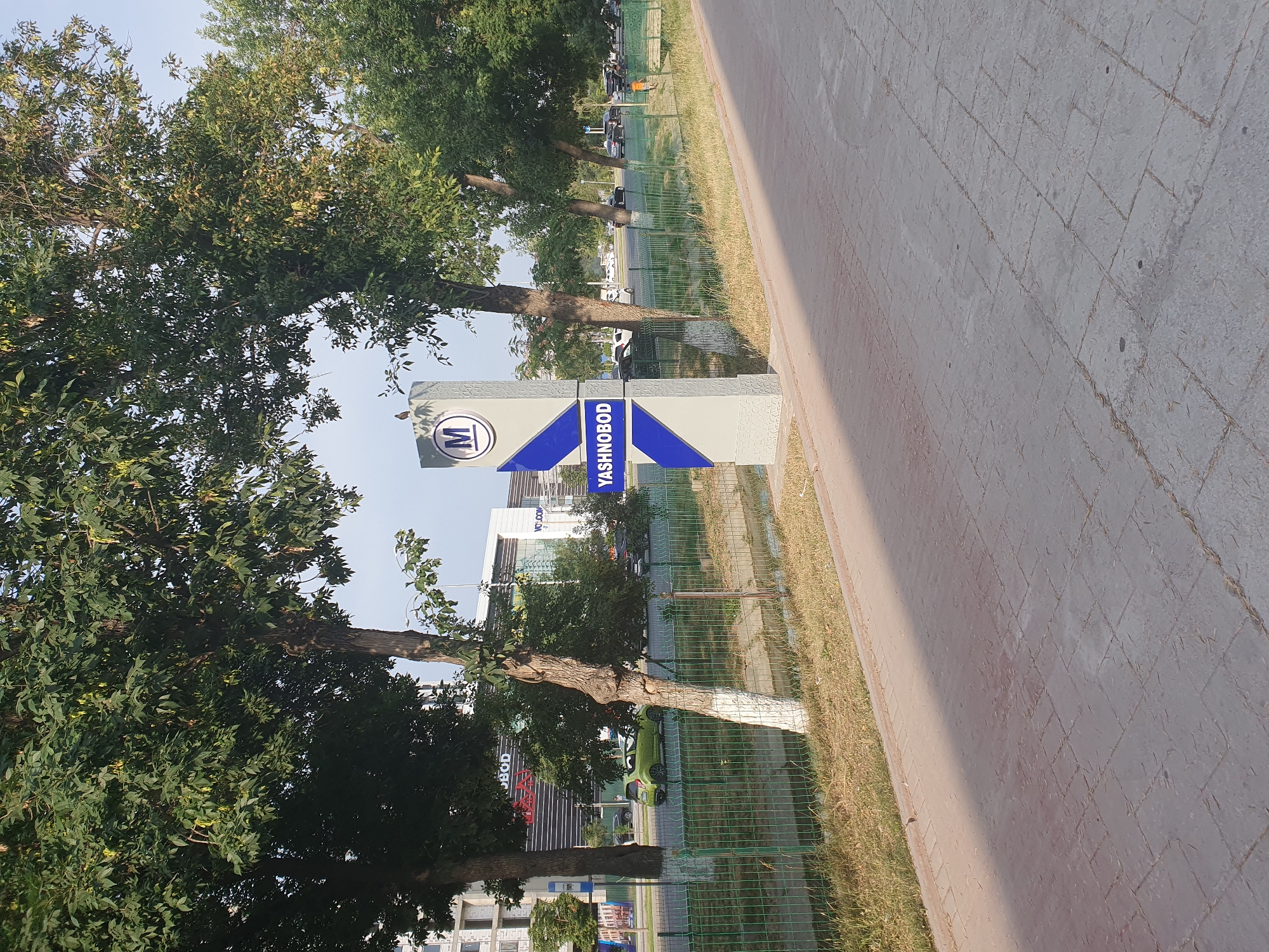The image shows a metro station sign that reads "YASHNOBOD" in Tashkent, Uzbekistan. The sign is a tall, grey pylon with blue accents and a circular "M" logo at the top. A bird is perched on the sign. In the background, there are trees, a street with cars, and modern buildings. The scene is set on a sunny day, with the sun casting a shadow across the paved walkway in the foreground.