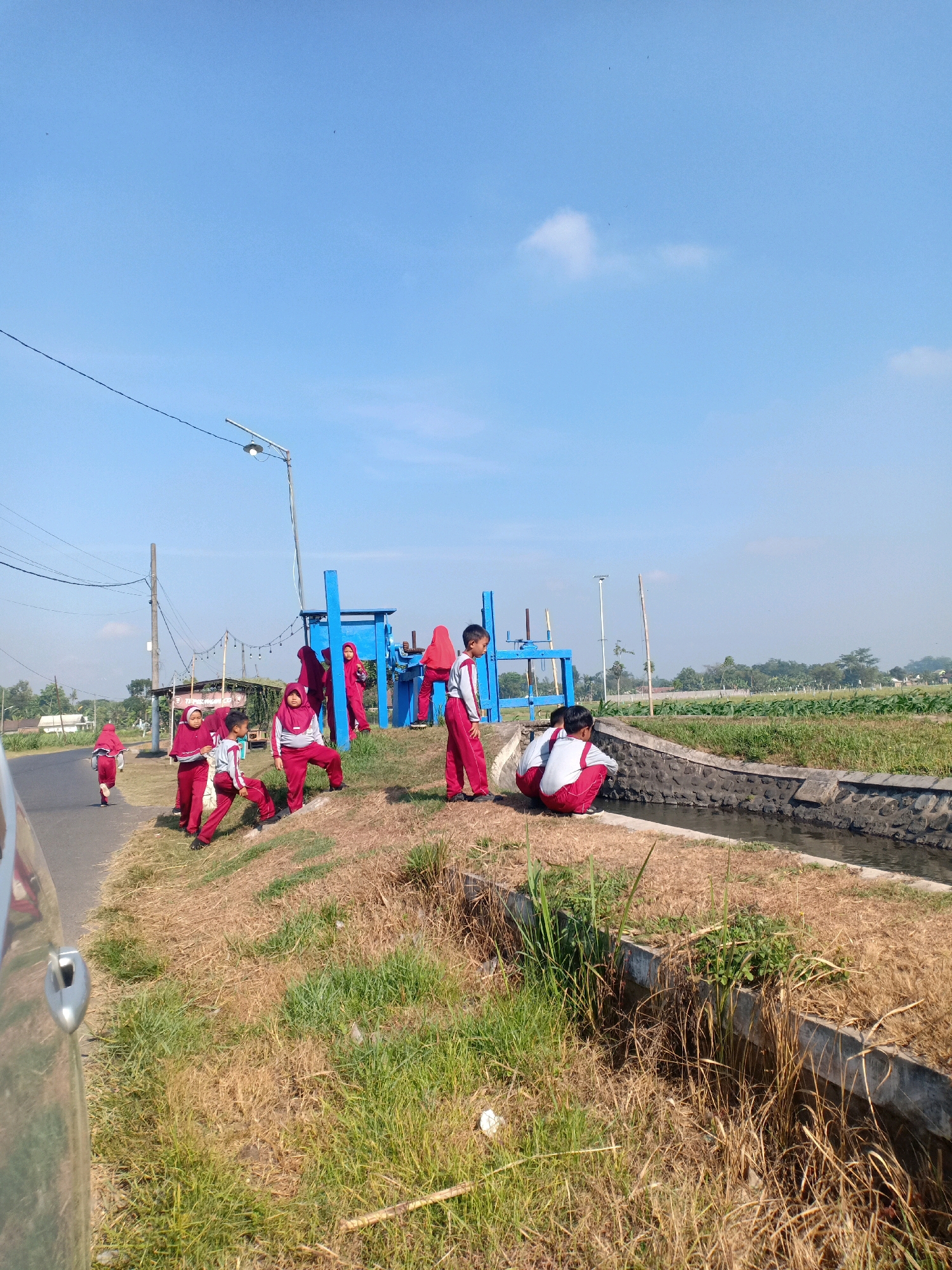 The image depicts a group of children in red and white uniforms near a blue irrigation gate in Kebonsari, Indonesia. The scene is set outdoors on a sunny day, with a clear blue sky. A paved road runs along the left side of the image, bordered by dry grass and vegetation. A ditch with water runs alongside a stone wall on the right. Several children are engaged in various activities: some are standing and observing, while others are sitting by the ditch. One child is running on the road. In the background, there are trees, agricultural fields, and residential structures, suggesting a rural or semi-rural environment. A street lamp is visible on a pole. The overall impression is of a school outing or a group of children exploring their surroundings.