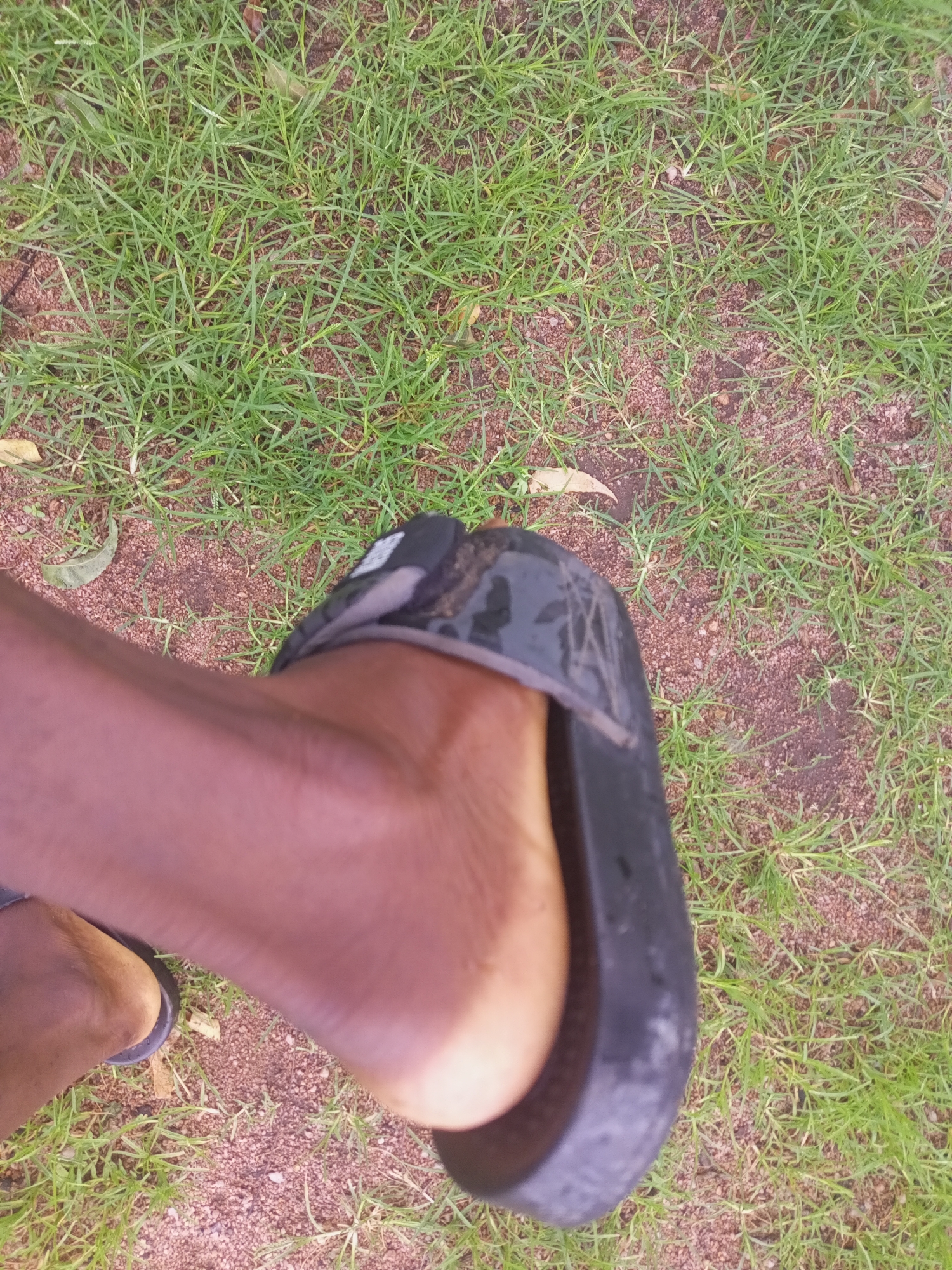 A close-up, top-down shot shows a person's bare foot wearing a black sandal, positioned on an area covered with sparse green grass and reddish-brown soil. The sandal appears to be made of rubber or a similar material, with a textured sole visible. A portion of the person's leg, which is tanned, is also in the frame. The background consists of more grass and soil. This scene is observed in Zinna, Nigeria.