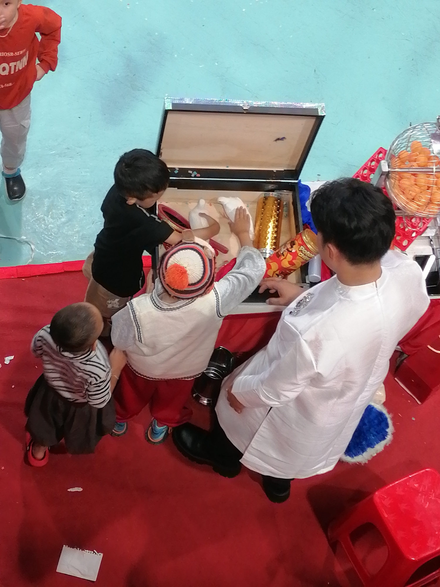 In Da Lat, Vietnam, a group of children and an adult are gathered around an open wooden chest on a red carpet. The chest contains various items, including a white cloth, a golden cylindrical object, and what appears to be a bag of chips. One child, wearing a red shirt and light-colored pants, stands to the left, looking down. Another child in a black shirt is examining the contents of the chest. A third child, in a white vest and red pants, is also looking into the chest, wearing a distinctive hat with red and white stripes and an orange pom-pom. A fourth child, the youngest, wearing a striped shirt and dark skirt, stands to the left, looking towards the chest. An adult in a white robe stands on the right, observing the scene. The background shows a light blue surface, likely a floor or wall, and a wire basket filled with orange items, possibly ping pong balls or snacks. The overall scene suggests a playful or educational activity.