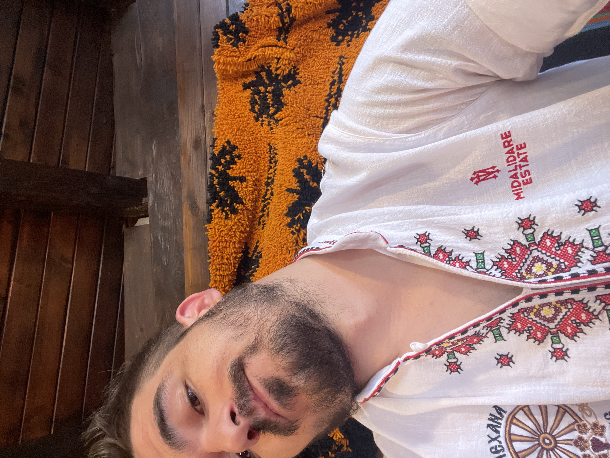 A man with dark hair and a beard is looking at the camera, his head tilted slightly to the side. He is wearing a white shirt with red and green embroidery. On the left side of his shirt, a wheel symbol is visible, and on the right side, the words "MIDALIDARE ESTATE" are embroidered in red. He is lying down, with an orange and black patterned blanket behind him. The background appears to be a wooden ceiling with beams. The location is Unknown, Unknown.
