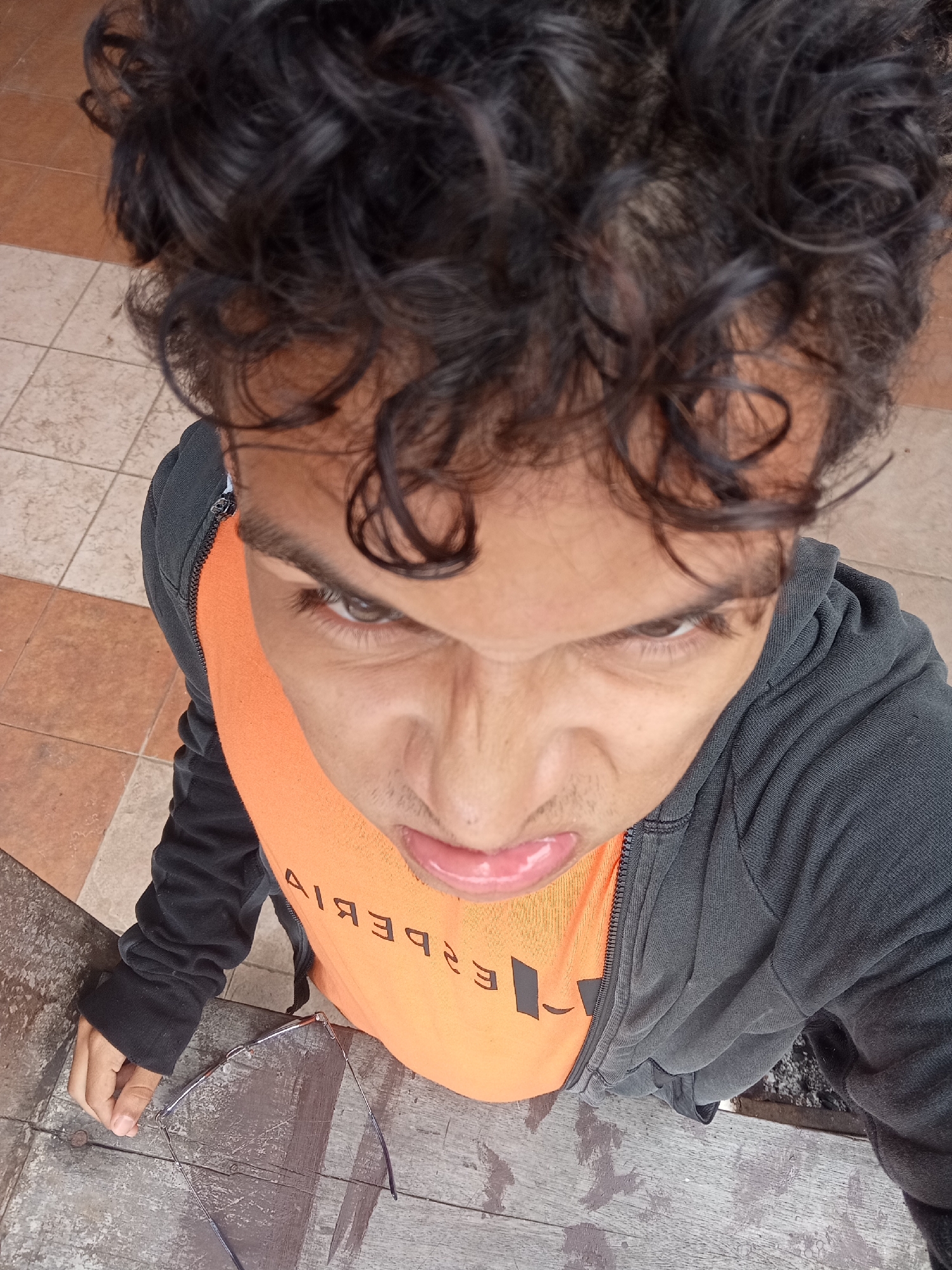 A high-angle shot shows a person with curly dark hair wearing an orange t-shirt under a gray zip-up hoodie. The person is sticking out their tongue and squinting. A pair of eyeglasses sits on a wooden surface below the person. The background shows tiled flooring. The location is Playa El Agua, Venezuela.