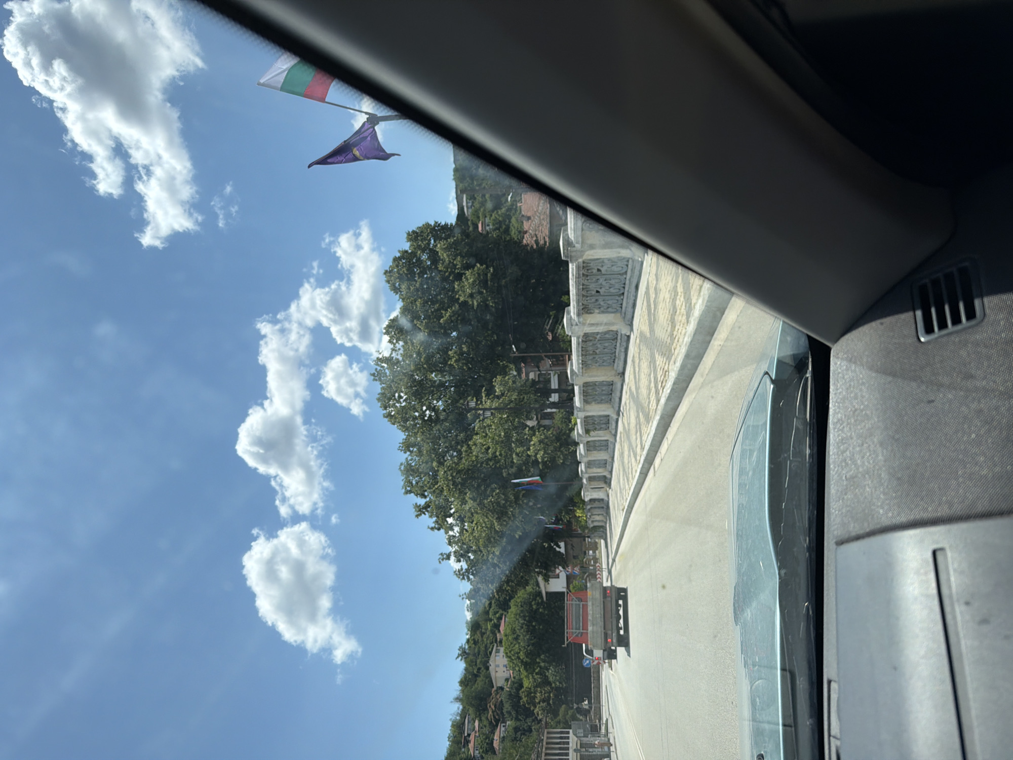 The image captures a view from inside a vehicle, looking out towards a street in Lovech, Bulgaria. The road leads towards a bridge adorned with decorative railings. On the bridge, two flags are visible, one predominantly purple and the other a tricolor of white, green, and red, likely the Bulgarian national flag. Beyond the bridge, a red truck is driving away from the camera on the road. The background reveals a hillside covered with houses and lush green trees, under a bright blue sky with scattered white clouds. The interior of the car shows the dashboard and windshield framing the external scene.