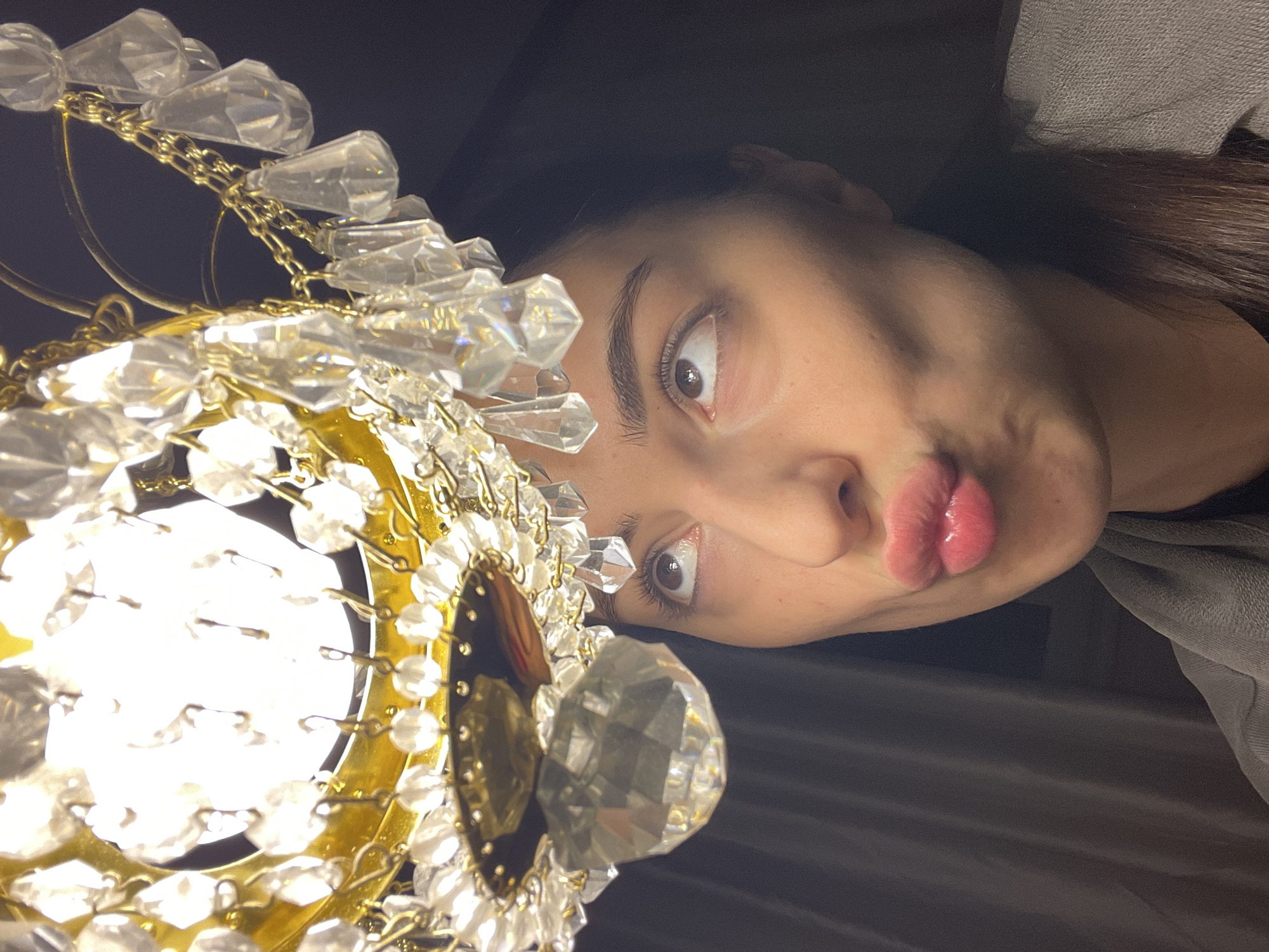 A dimly lit, indoor scene depicts a female subject looking upwards at a brightly illuminated chandelier. The chandelier, occupying the upper frame, features a gold-toned metallic structure adorned with numerous clear, multi-faceted crystal pendants and beads. A strong central light source within the chandelier creates a bright glow. The female, positioned below, is shown from the chest up, with her head slightly tilted back and her dark eyes rolled upwards. Her lips are pursed in a "kissing" expression, with visible pink lip gloss. She wears a grey garment over a dark top. The background is dark and indistinct, with a dark, textured vertical surface visible to the left. The specific location within Moscow, Russia, cannot be determined from the visual information provided.
