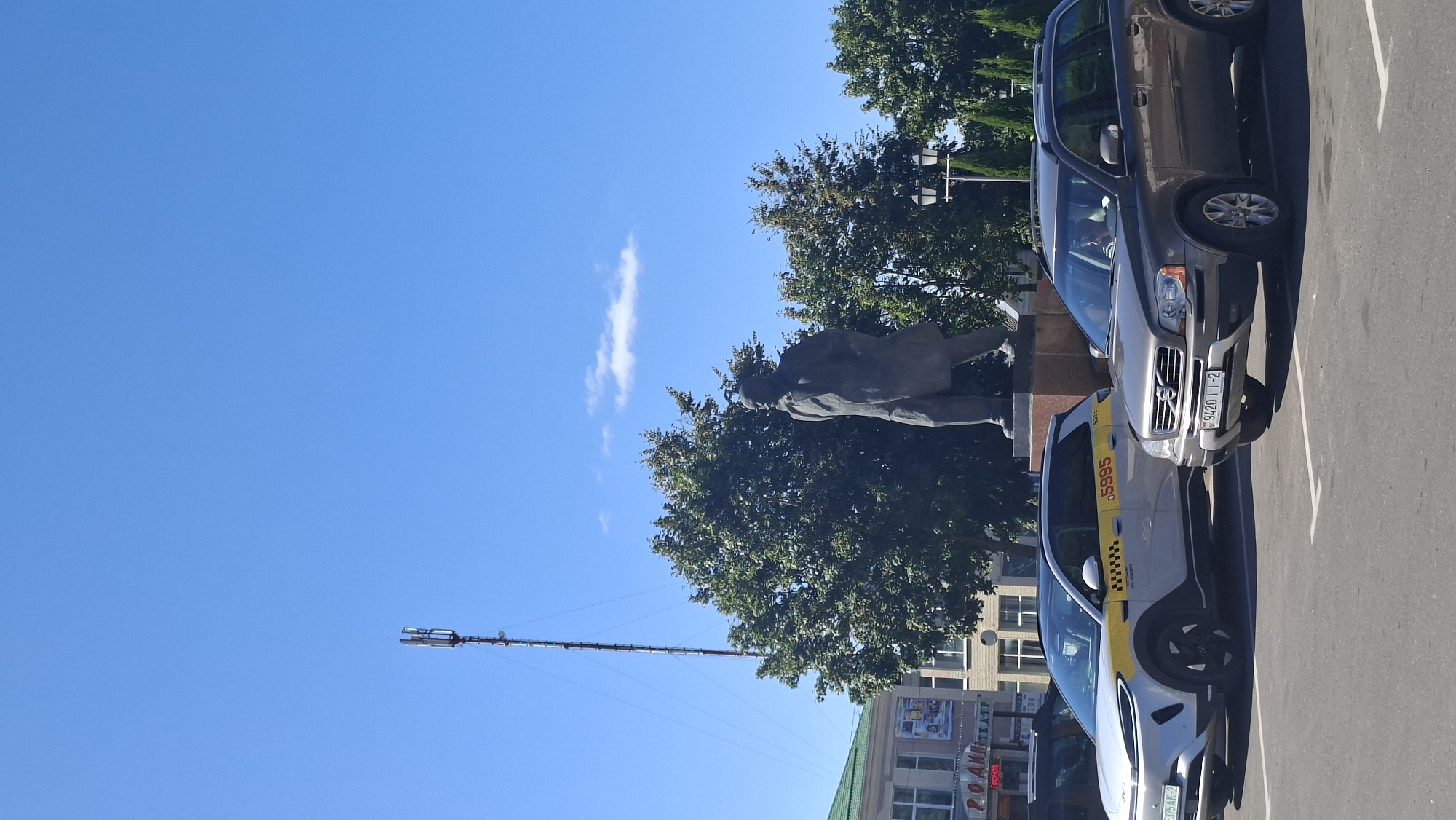 The image depicts a sunlit outdoor scene in the city of Pastavy, Belarus. In the foreground, two cars are parked on asphalt. The car on the left is a white SUV with yellow and black taxi markings, displaying the number "5995". The car on the right is a gray Volvo SUV with a Belarusian license plate reading "9420 II-2". Behind the cars, a bronze statue of a man in a coat stands on a pedestal. Tall green trees form a backdrop, with a telecommunications tower visible in the distance on the left. A building with a green roof and signage for "РОДИНА ТЕАТР" (Rodina Theater) is partially visible on the far left. The sky is a clear blue with a few scattered white clouds.
