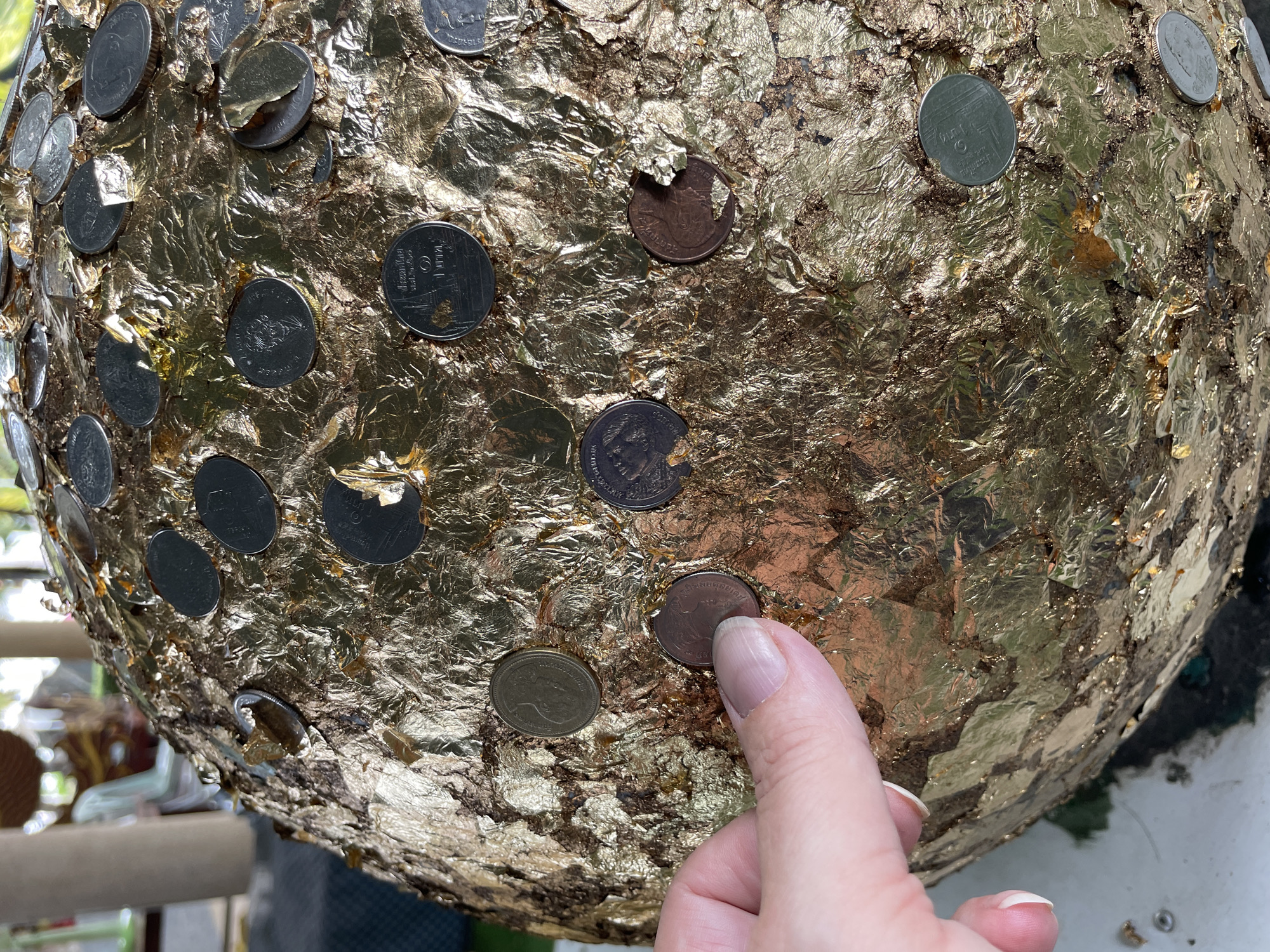 A close-up, eye-level shot captures a hand reaching out to touch a large, spherical object adorned with numerous gold-leafed coins. The object is covered in crinkled gold foil, with coins of various sizes and denominations pressed into its surface. Some coins are visible with Thai inscriptions and imagery. The hand, with manicured fingernails, is positioned in the lower left quadrant of the frame, its index finger gently pressing against one of the coins. The background is blurred, suggesting an outdoor setting, possibly a temple or sacred site in Tambon Na Chom Thian, Thailand, given the cultural context of such offerings. The lighting is bright, highlighting the reflective surfaces of the gold foil and coins.