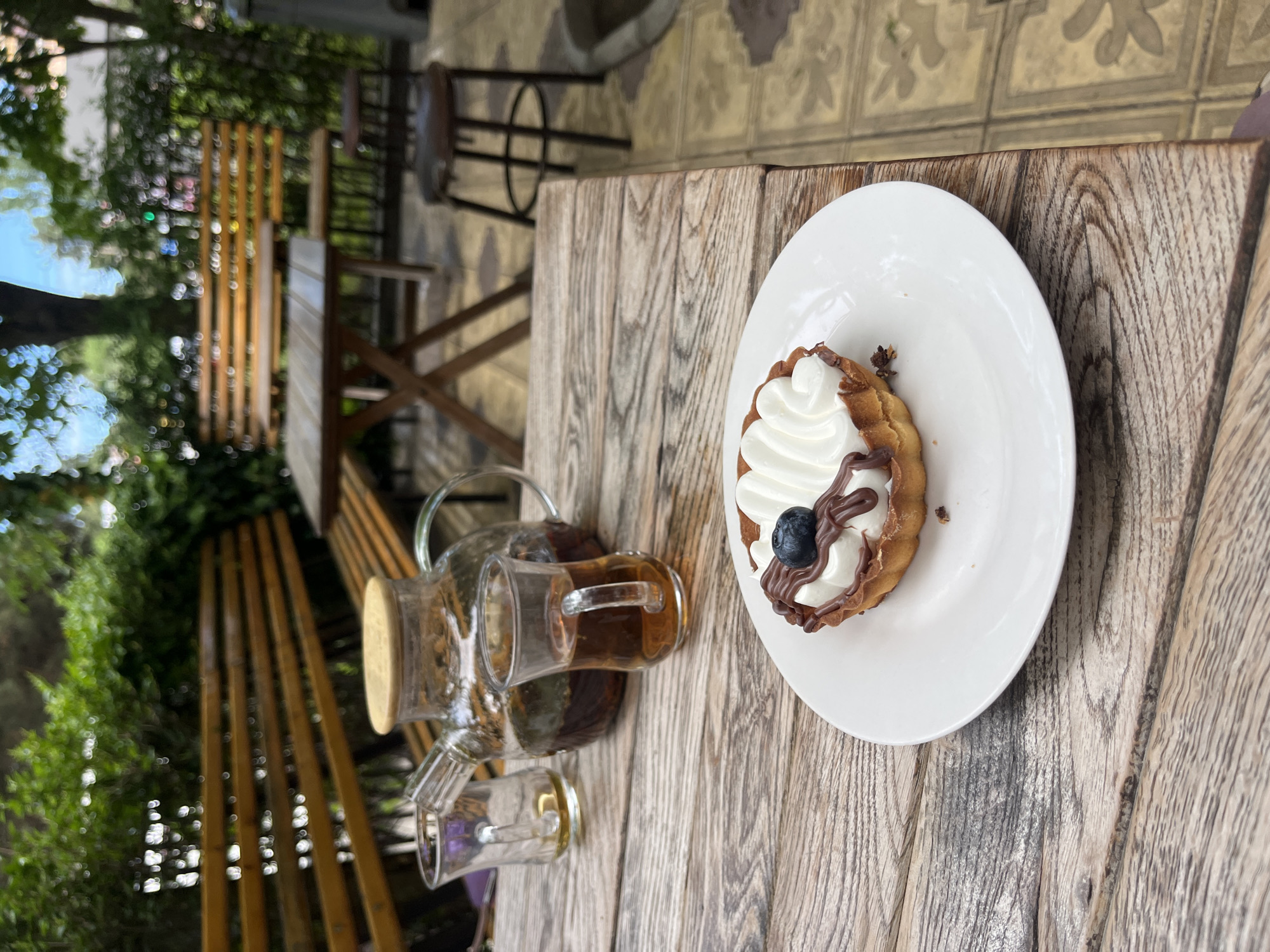On a weathered wooden table outdoors in Kaspiysk, Russia, sits a white plate with a tart topped with white cream, chocolate drizzle, and a blueberry. A glass teapot and two glass cups containing a dark liquid, possibly tea, are also on the table. In the background are benches, chairs, greenery, and other elements suggesting an outdoor café or patio setting. The image is rotated 90 degrees counter-clockwise.