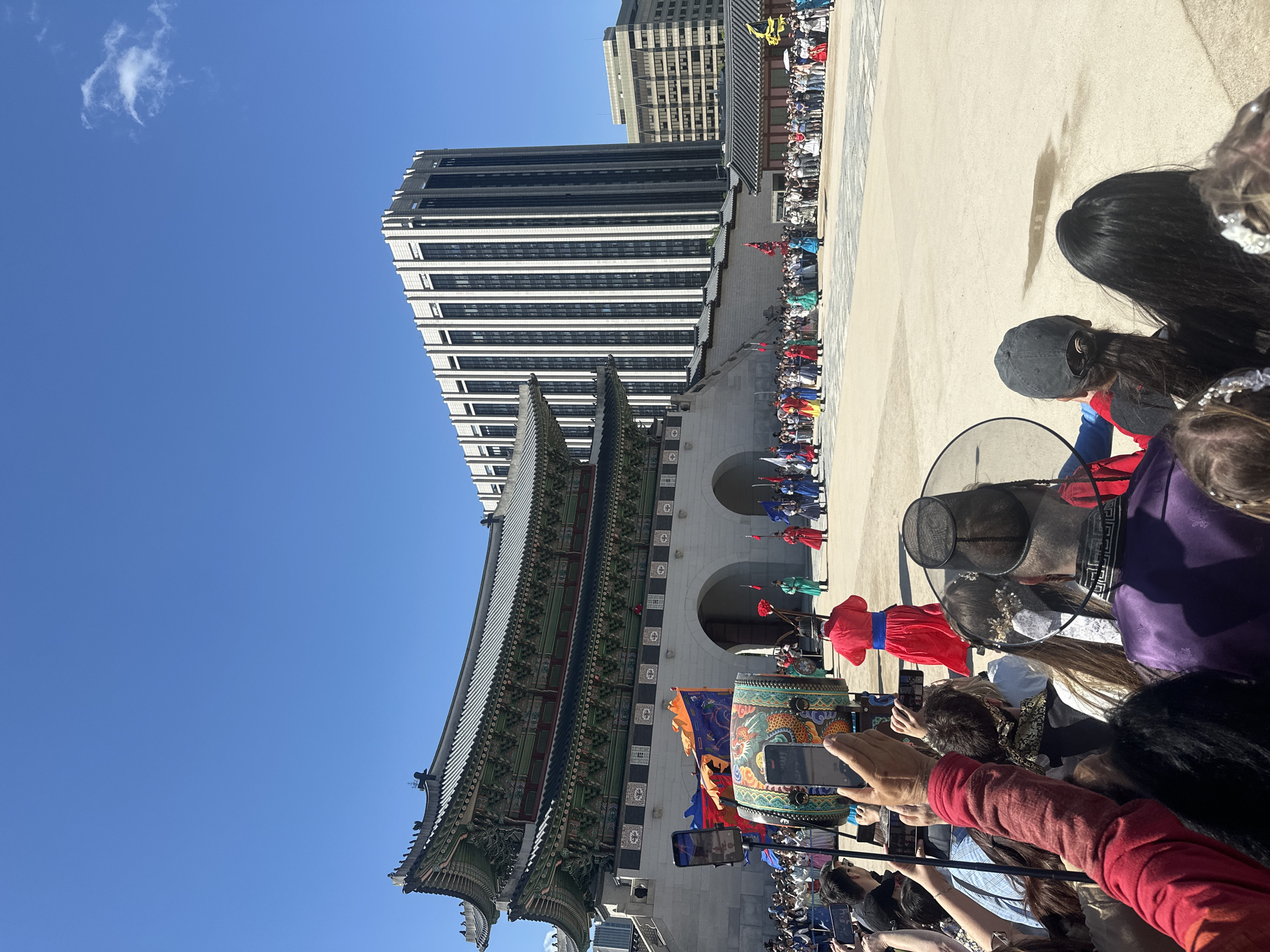 The image shows a traditional Korean royal guard changing ceremony at Gyeongbokgung Palace in Seoul, South Korea. A large crowd of people, many holding up their phones to take pictures or videos, are observing the ceremony. The ceremonial guards are dressed in colorful traditional Korean clothing. A large drum decorated with an ornate dragon design is visible. The gate of the palace is visible as well as a tall, modern building. The sky is a clear blue.