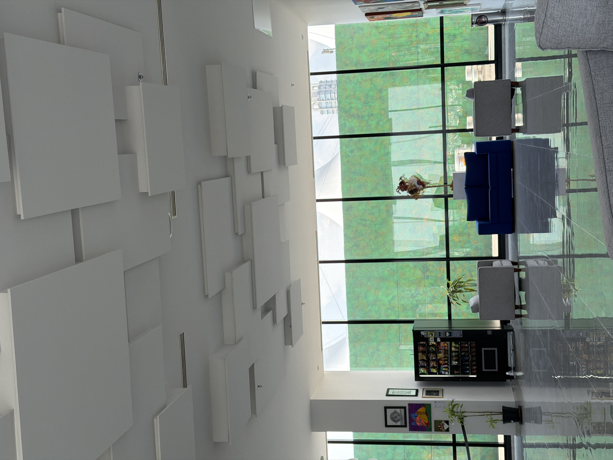 The image shows the interior of a modern building in San Bernardino Tlaxcalancingo, Mexico. A white wall with square, white panels in a geometric design dominates the left side of the image. Large windows span the width of the scene, revealing an exterior view of greenery. A blue sofa and gray chairs provide seating. A vending machine stands in the corner. Artwork and plants are displayed along the wall. The floor is polished and reflective.