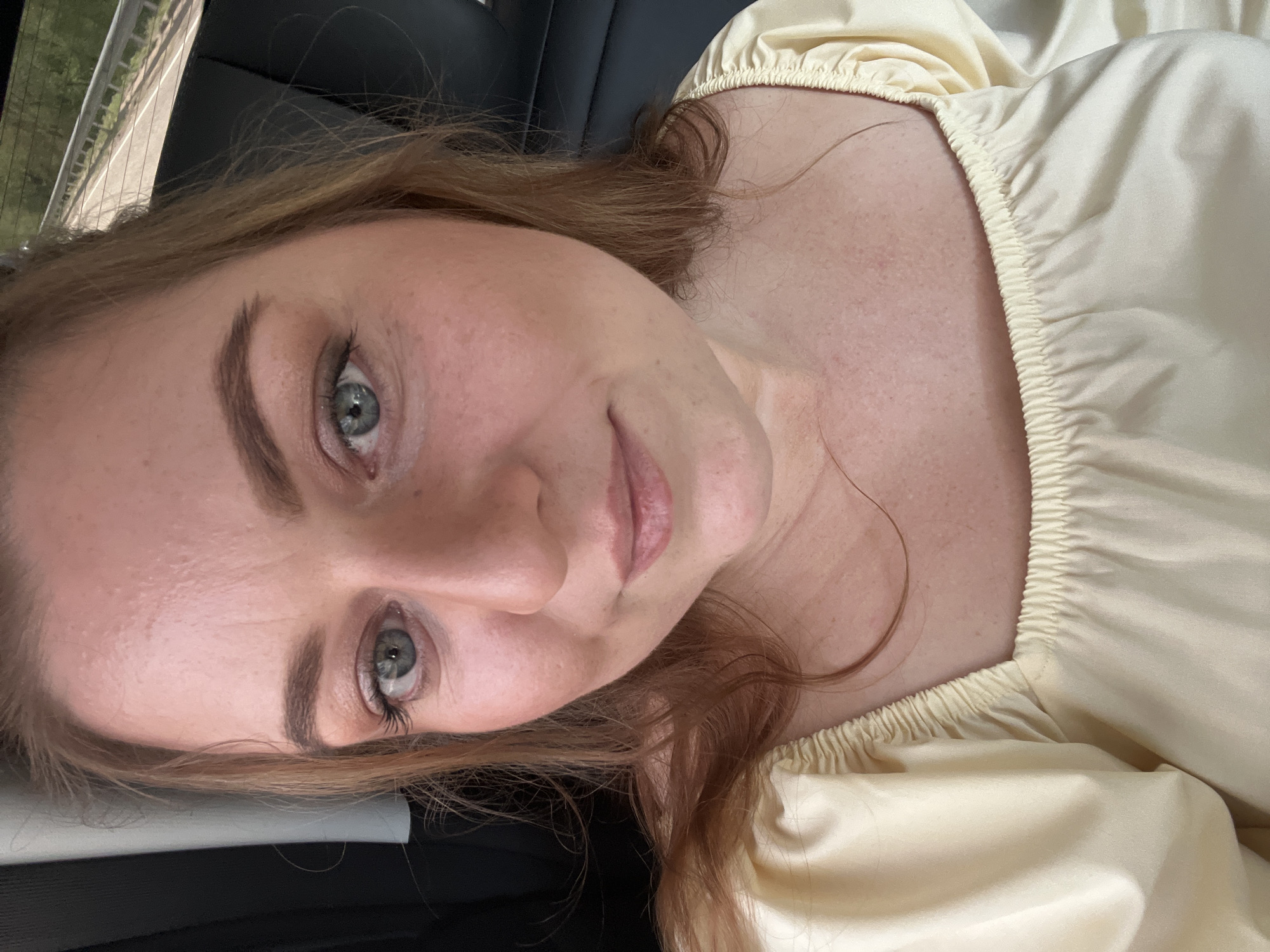 The image is a selfie of a fair-skinned woman with light brown hair and grey eyes. She is wearing a light yellow, possibly satin or silk, top with a gathered neckline. Her face is partially obscured by the perspective, but visible features include prominent eyebrows, freckles, and a slightly downturned mouth. The background is dark and suggests the interior of a car, possibly a leather seat. Out of focus, in the very upper left corner, what appears to be either landscape or a building can be seen. There is no information in the image to determine if this photo was taken in or near Sin'kovo, Russia.