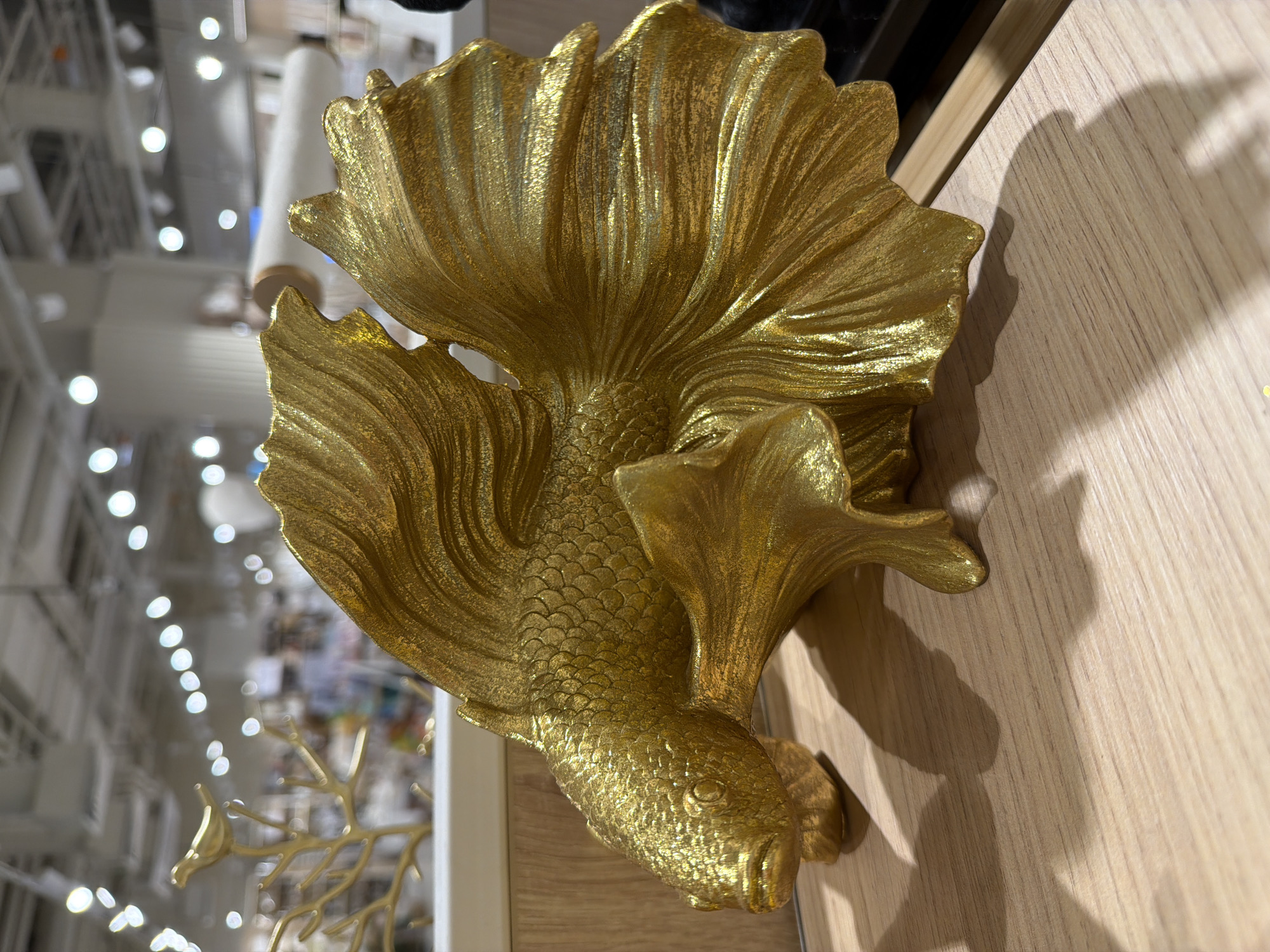 A close-up shot features a golden betta fish statue placed on a light-colored wooden surface. The statue is highly detailed, showing the fish's scales and flowing fins. The location is unknown, and the photograph is taken indoors under artificial lighting, casting shadows onto the wooden surface.