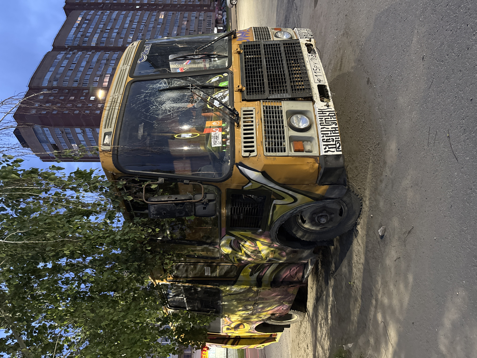 An abandoned, graffiti-covered yellow bus with a cracked windshield sits on a dirt and asphalt lot at dusk in Ulyanovsk, Russia. The bus is partially obscured by a tree with green foliage. The bus is old and appears to be in disrepair, with rust visible in several areas. The graffiti on the side of the bus is abstract and colorful. In the background, a modern, multi-story brick apartment building stands. The sky is a dusky blue. The ground is a mix of dirt, gravel, and asphalt, with shadows cast by the bus and the tree.