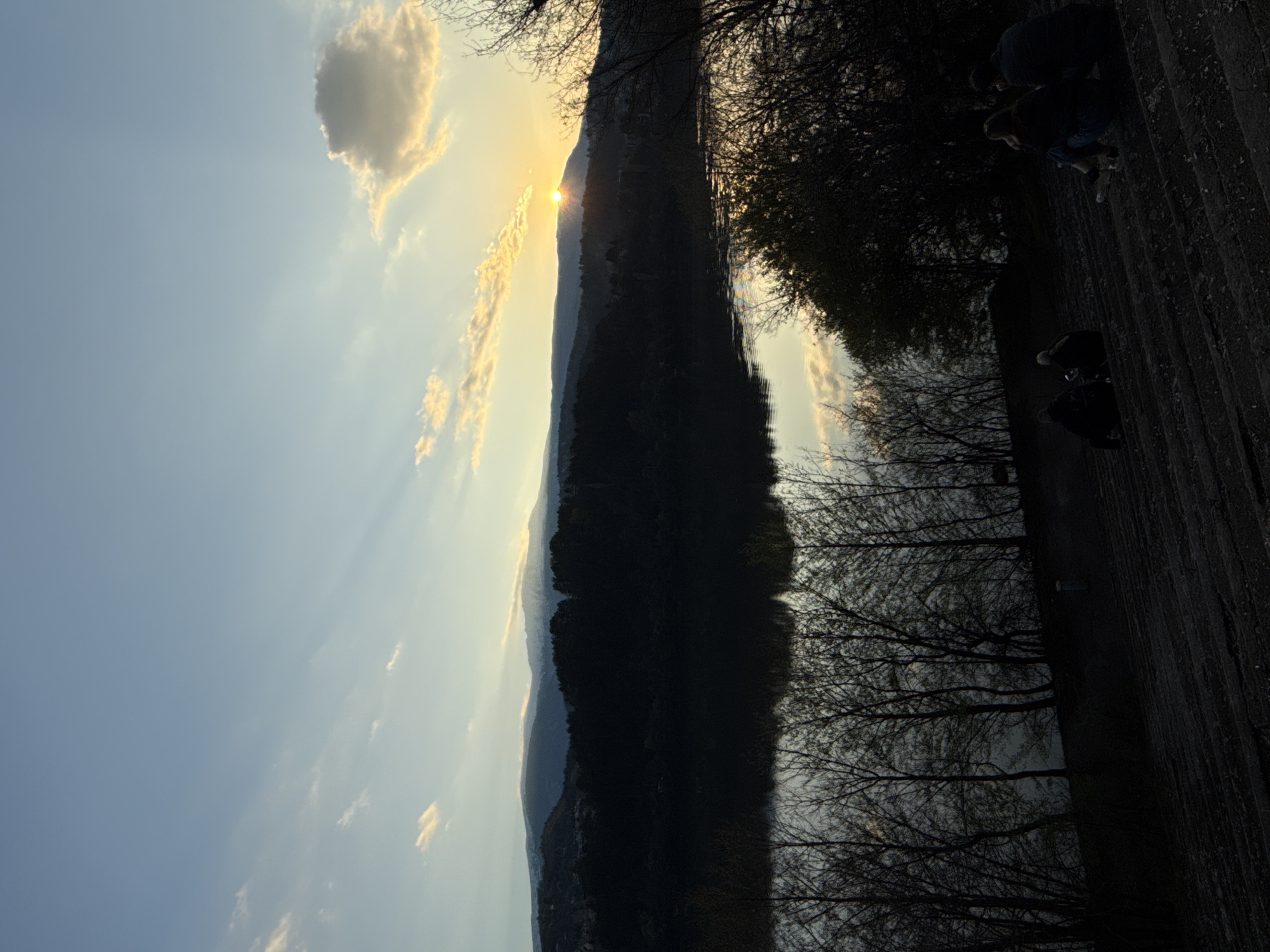 The image shows a landscape in Pancharevo, Bulgaria, at sunset. The sun is setting behind a mountain range, creating a silhouette. Light rays shine through the clouds. In the foreground, there are bare trees and people standing on what appears to be a dirt or wooden surface. The overall scene is dark.