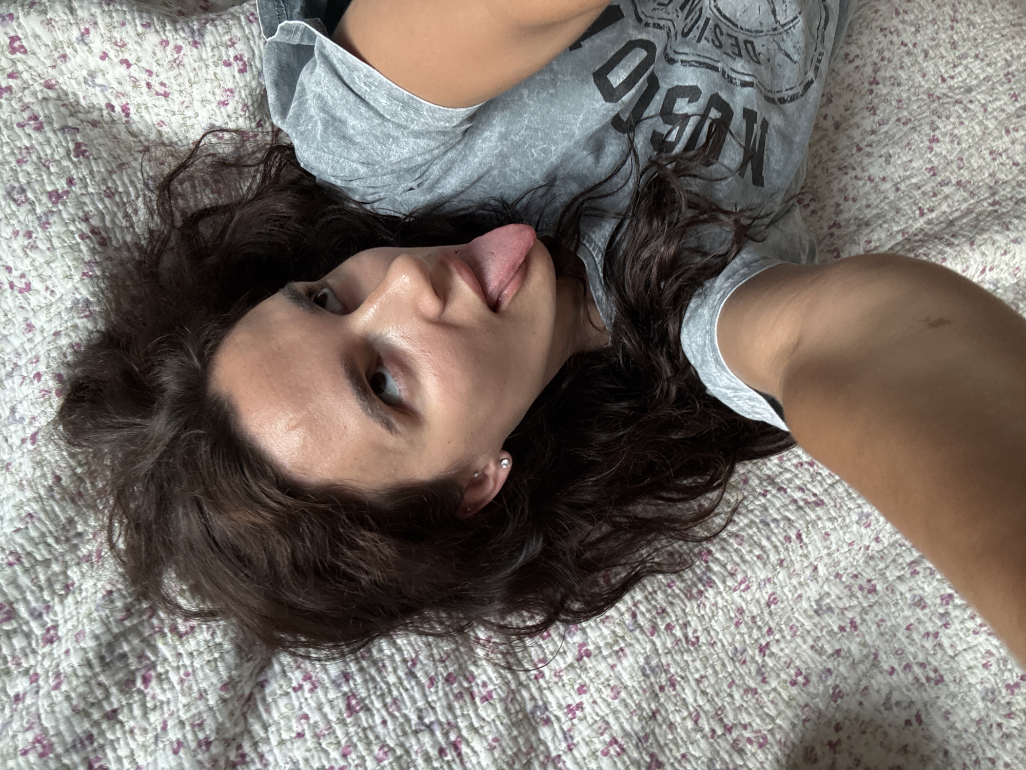 A young woman with dark brown, wavy hair is lying on her back on a floral patterned bedspread. She is wearing a grey t-shirt with black lettering. Her head is tilted back, and her eyes are looking upwards. Her tongue is sticking out to the side. The lighting suggests an indoor setting, possibly a bedroom. There are no visible landmarks or any indication of the city of Chaikovsky, Russia.