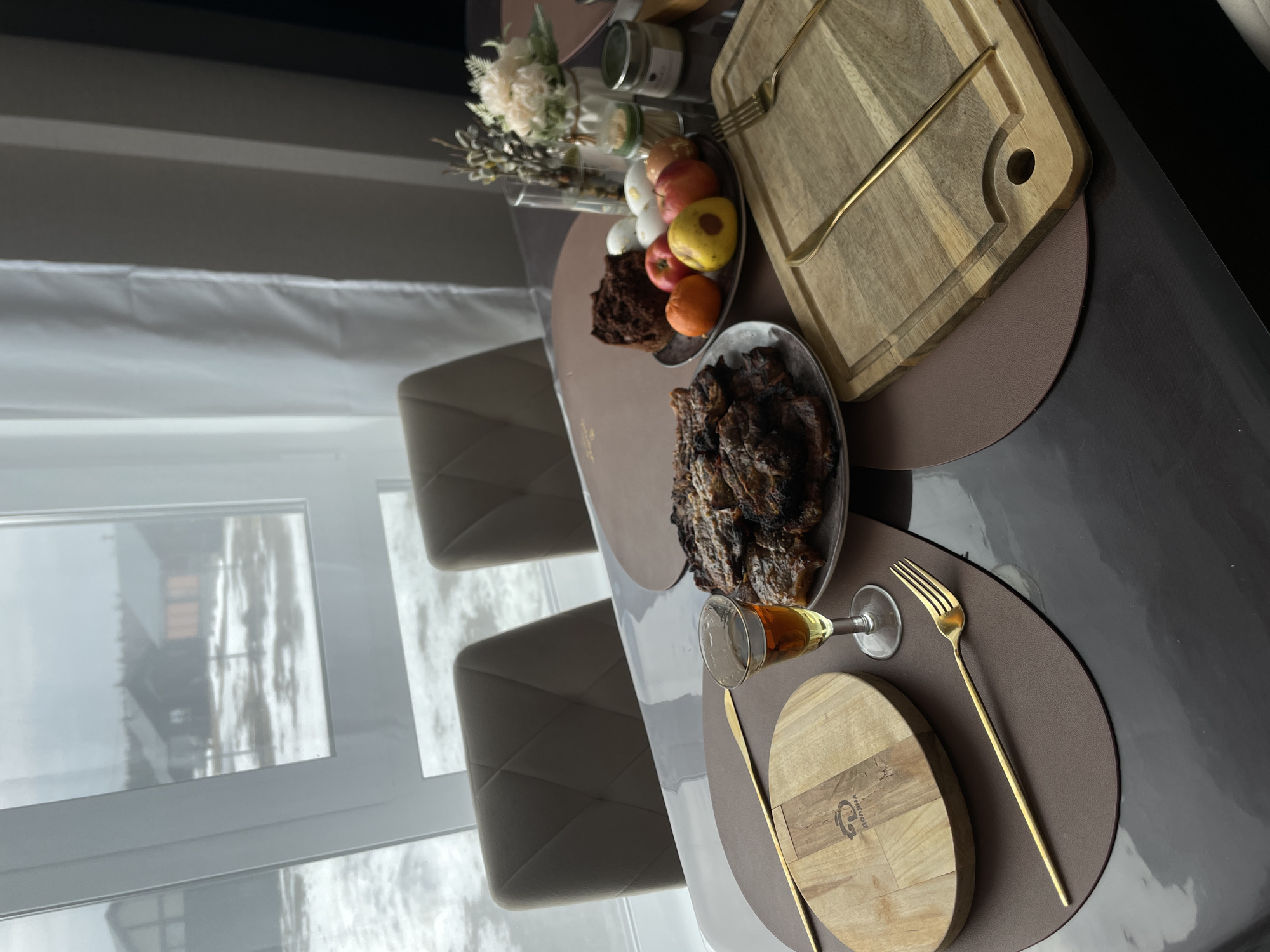 A table is set for a meal next to a window looking out onto a snowy landscape. The table is covered with brown placemats. On the placemats are a platter of roasted meat, a glass of amber liquid, a wooden board, and golden cutlery. There is also a platter of fruit and cake, and a vase of flowers. The location may be in or near the city of Подшивалово, Russia, given the snowy landscape visible through the window. The scene suggests a gathering or celebration.