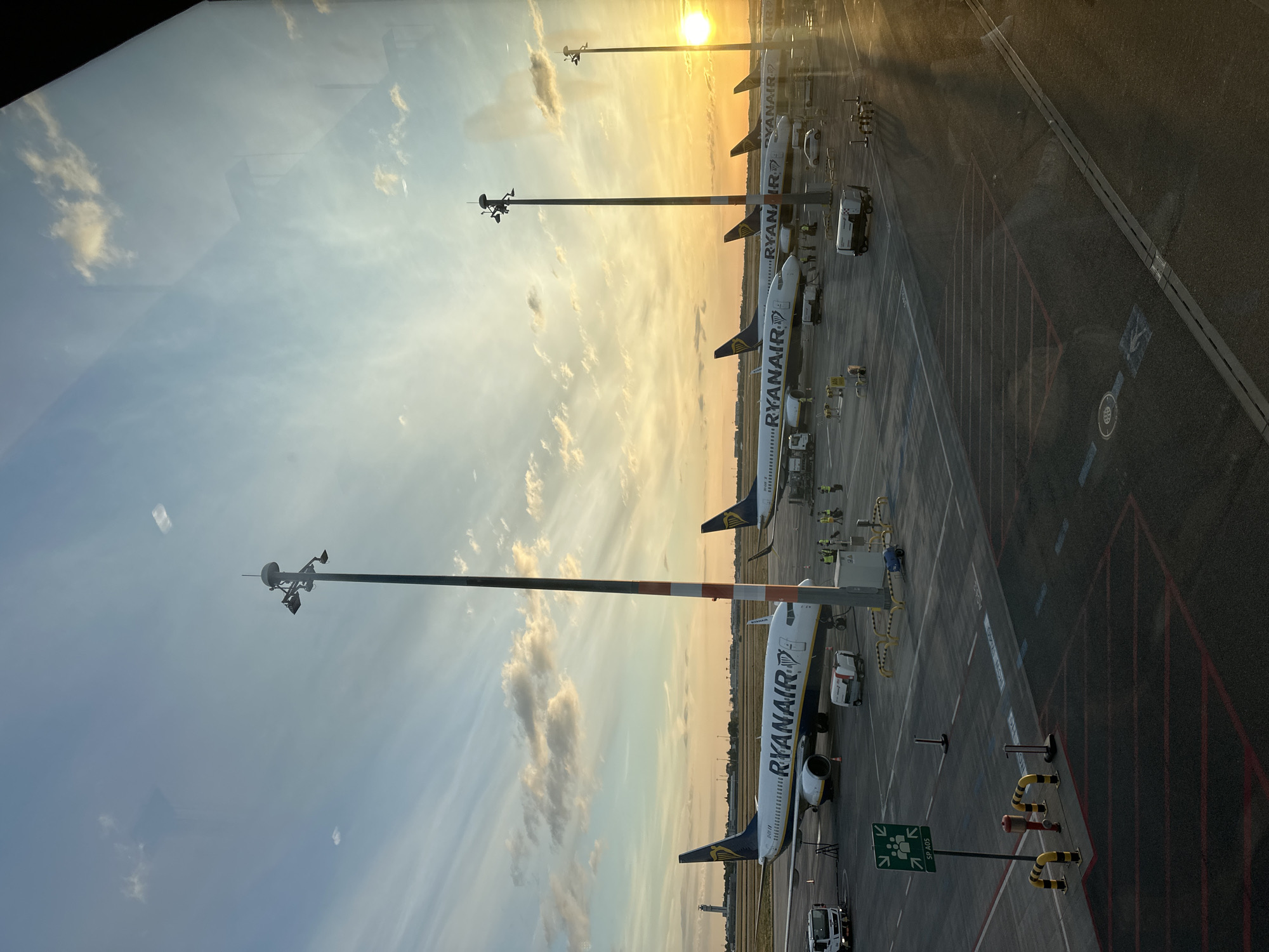 The image depicts an airport tarmac in Schönefeld, Germany, during a sunset. Several Ryanair airplanes are parked, with their logos clearly visible. Ground crew members in yellow vests are visible near the aircraft, appearing to be engaged in various tasks. Service vehicles, including a truck and smaller utility vehicles, are also present on the tarmac. The sky is a blend of warm sunset colors, with scattered clouds, casting a warm glow over the scene. Tall lighting poles are positioned throughout the tarmac. The overall impression is one of an active airport environment during the golden hour.
